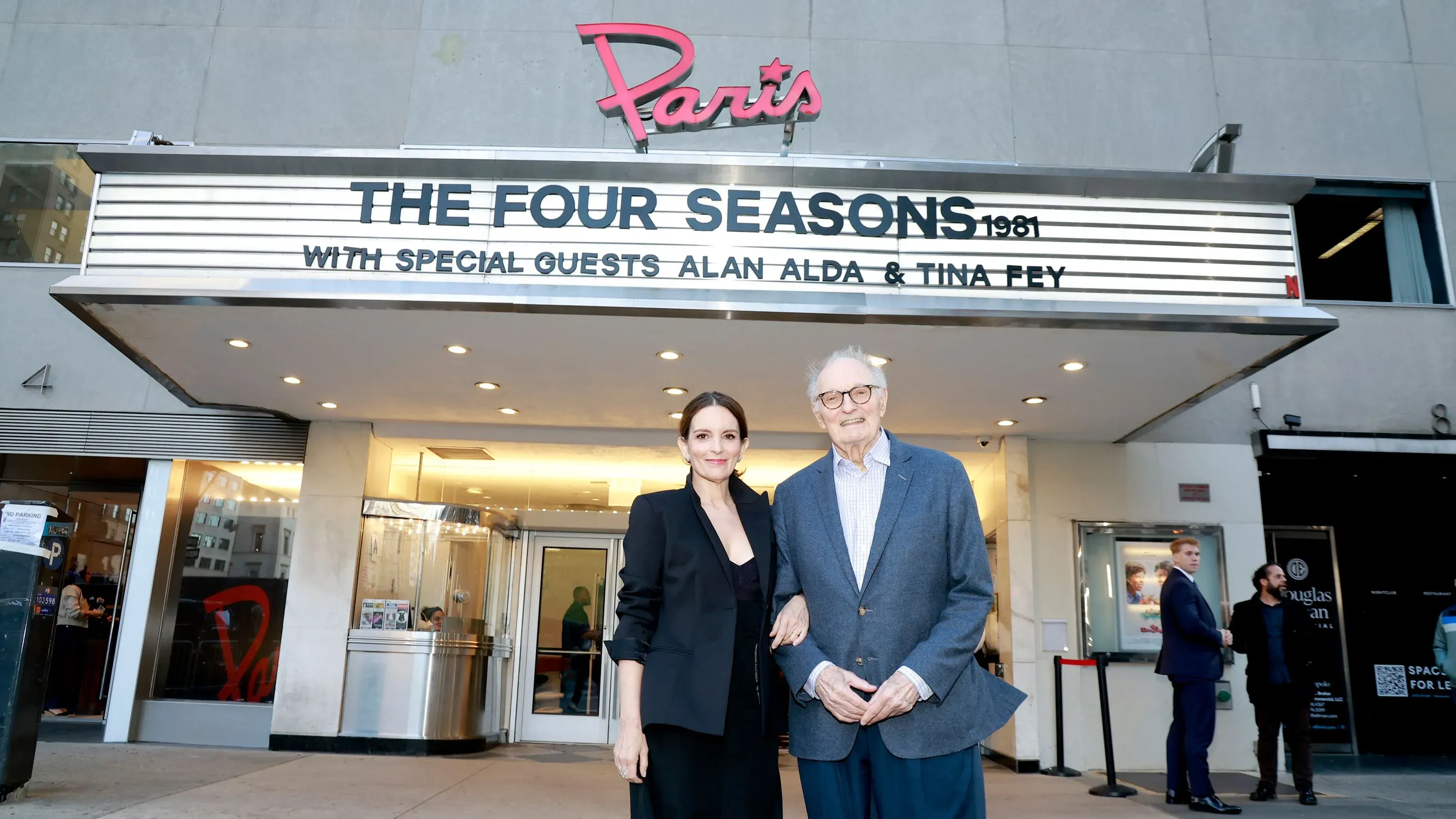 ألان ألدا وتينا فاي يقفان أمام مسرح "باريس" خلال عرض مسلسل "الفصول الأربعة" الجديد، مع لافتة ترويجية للفيلم الكلاسيكي.