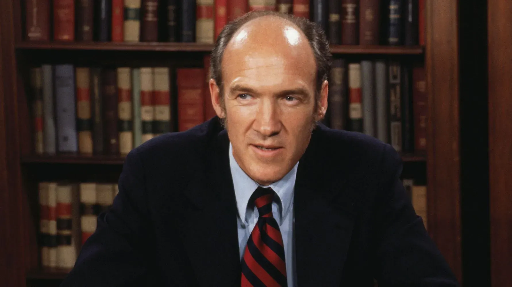 صورة لآلان سيمبسون، السيناتور الجمهوري السابق من وايومنغ، أثناء حديثه في مكتبه، مع خلفية من الكتب، يعكس شخصيته البارزة ومساهماته السياسية.