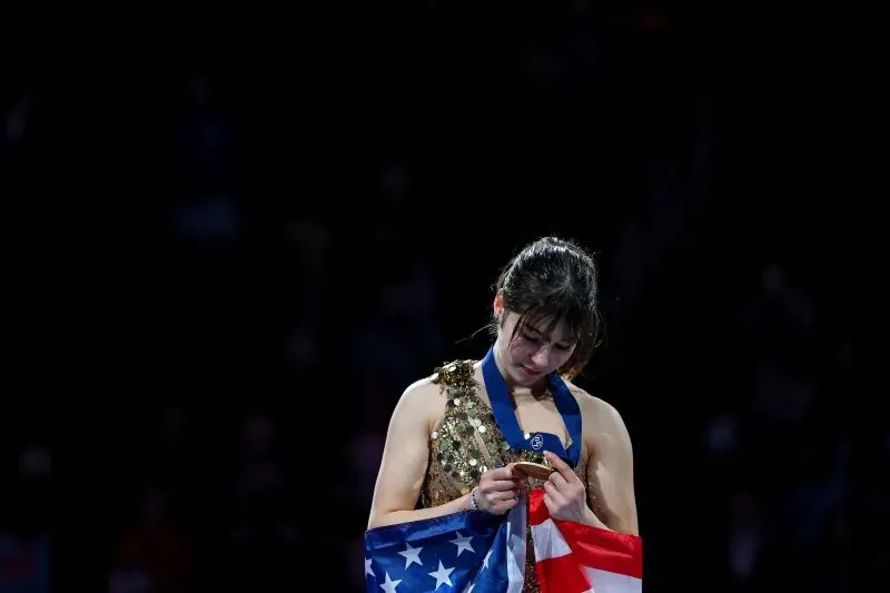 أليسا ليو، بطلة التزلج الأمريكية، تحمل ميداليتها الذهبية بفخر، بينما تنظر إلى العلم الأمريكي، تعبيرًا عن مشاعر الانتصار والامتنان.