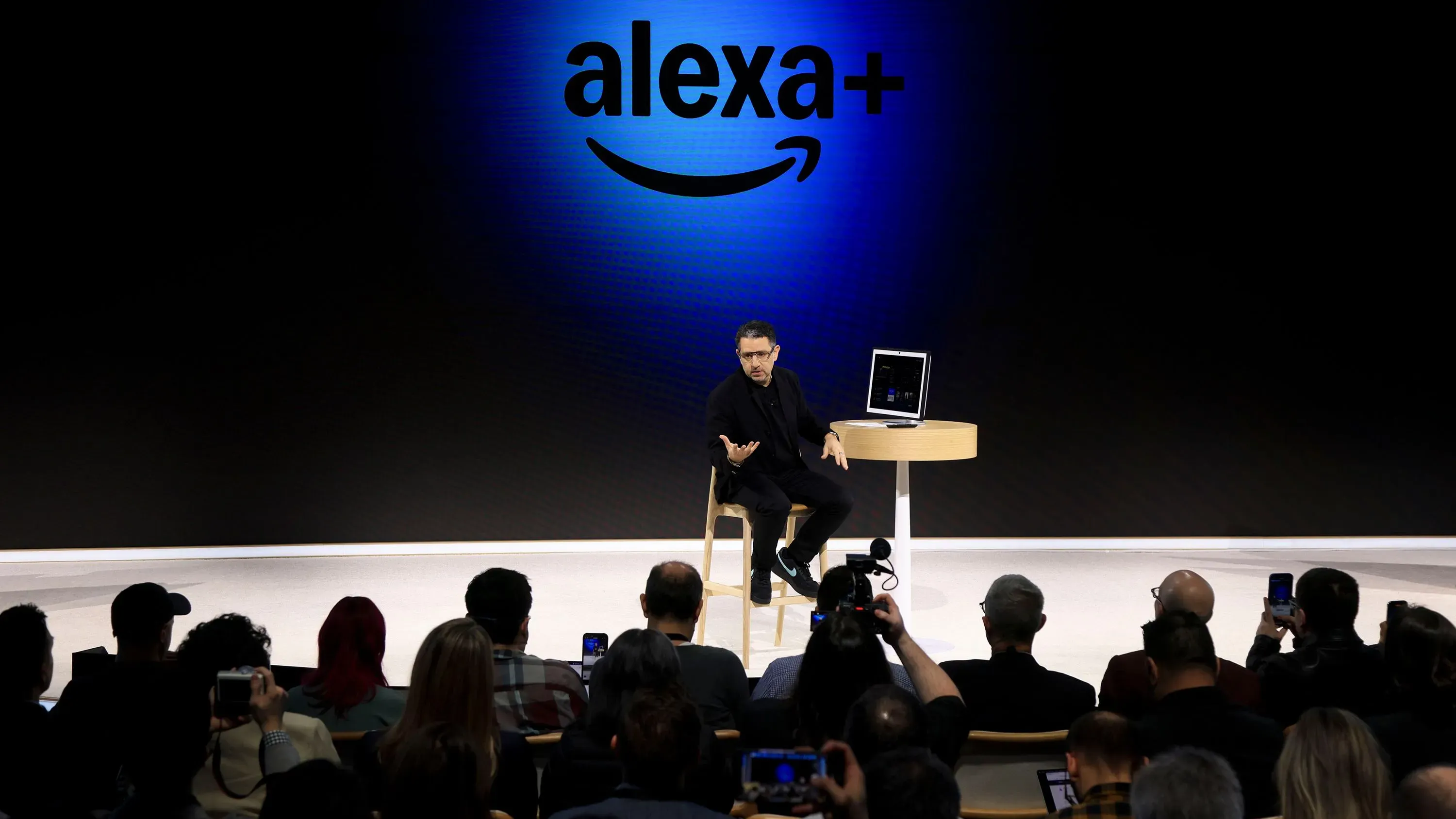 بانوس باناي يتحدث عن تحديثات أليكسا في حدث أمازون، مع جمهور يلتقط الصور خلفه، وشعار Alexa+ مضاء على الشاشة.