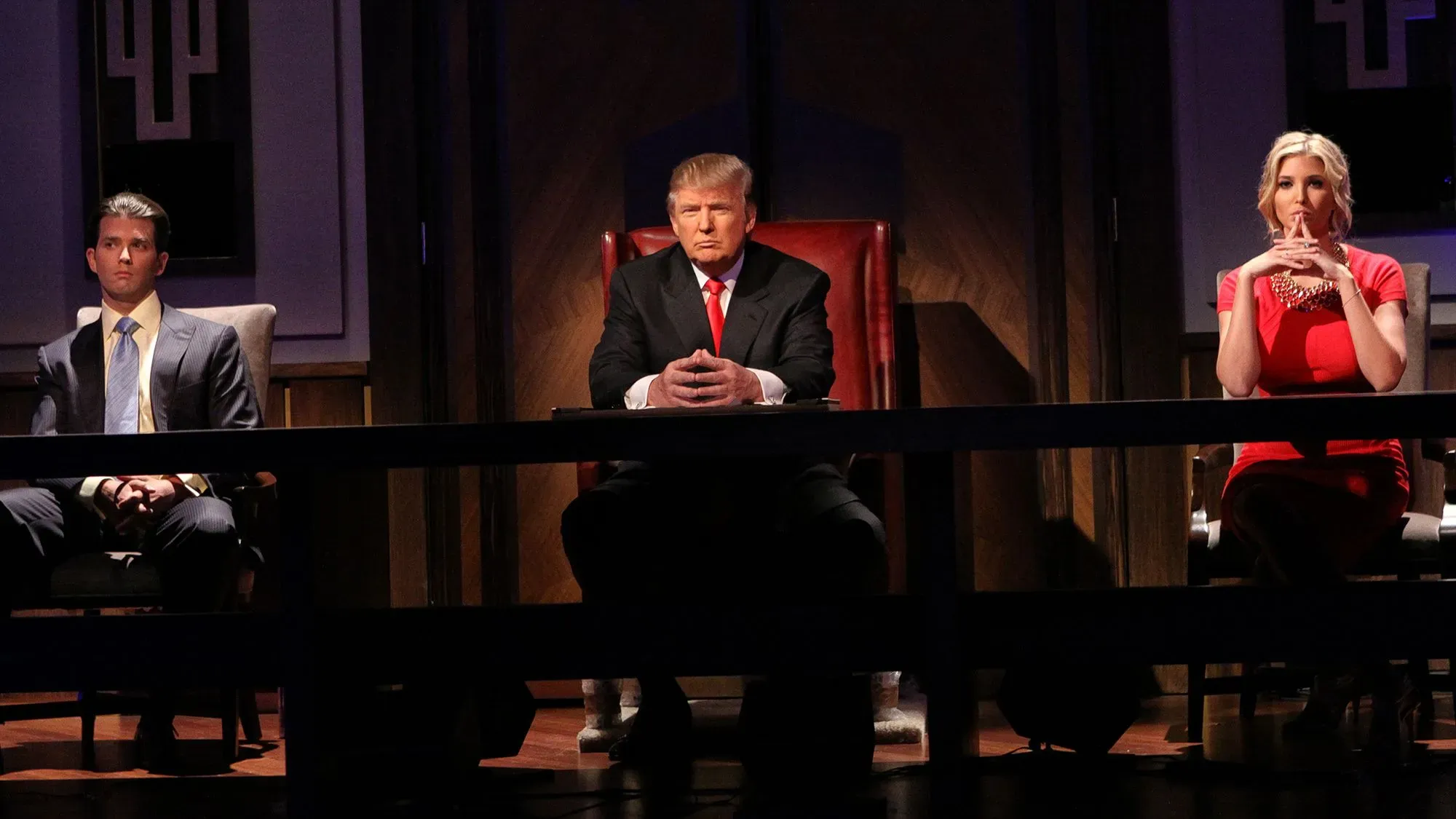 دونالد ترامب يجلس في الوسط خلال برنامج "The Apprentice" مع ابنه دونالد ترامب جونيور وابنته إيفانكا، في أجواء تنافسية.