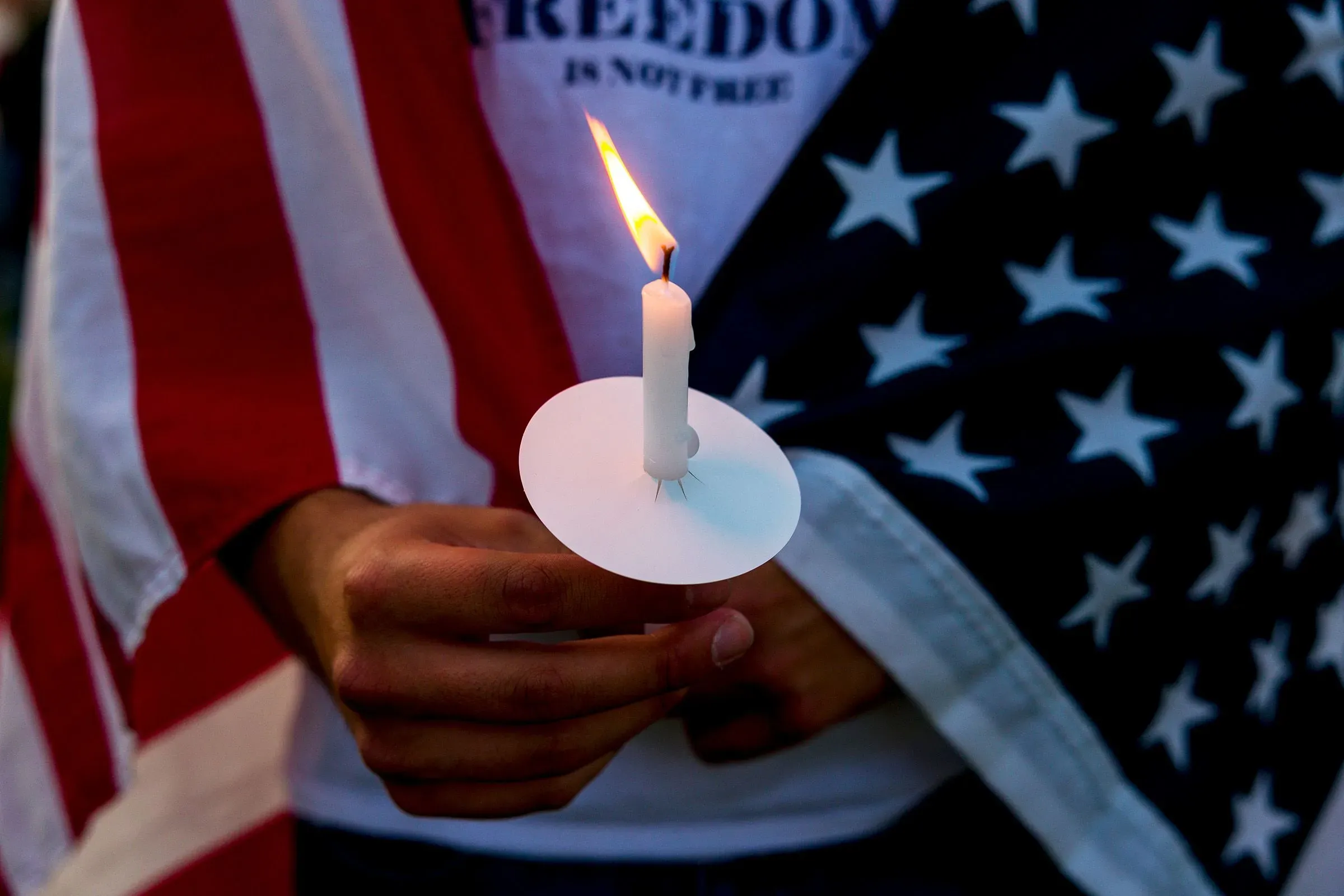 شخص يحمل شمعة مضاءة بينما يلتف حوله علم أمريكي، مما يعكس حالة الحزن والتأمل بعد اغتيال تشارلي كيرك وتأثيره على السياسة الأمريكية.