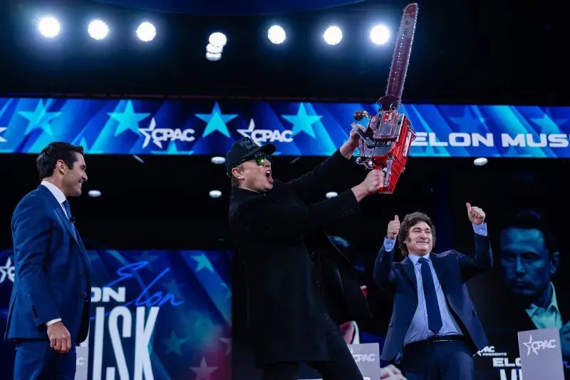 إيلون ماسك يحمل منشارًا كهربائيًا على المسرح في مؤتمر CPAC، مع جمهور متحمس، في مشهد يرمز لتحدي البيروقراطية الفيدرالية.