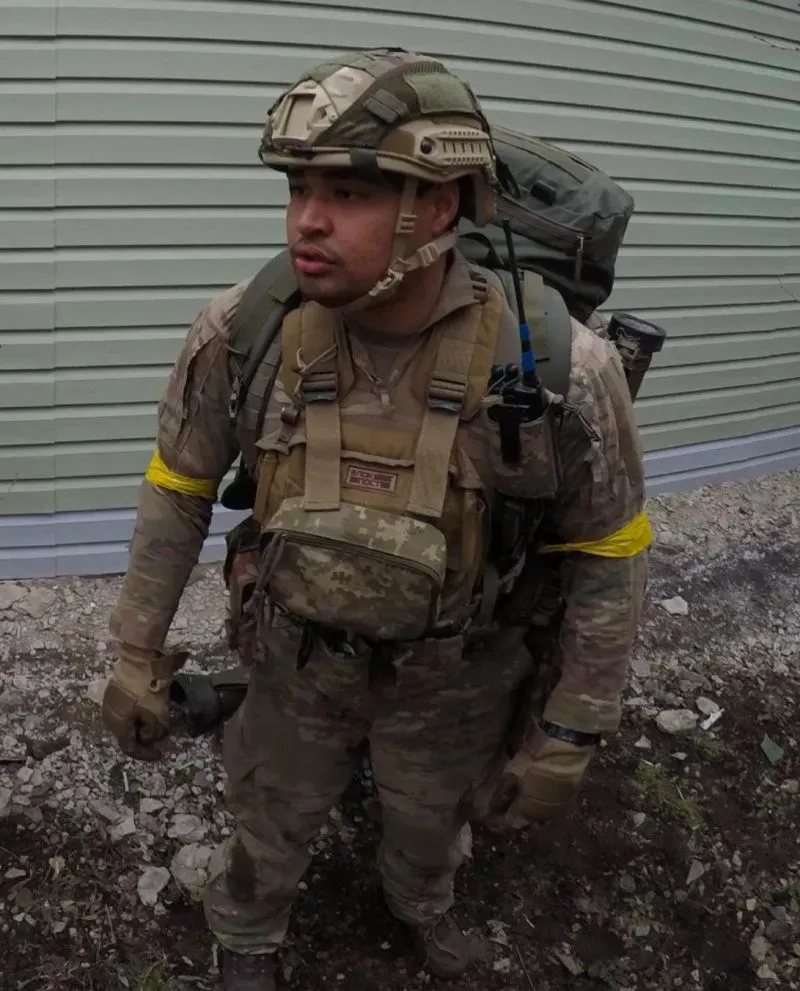 مقاتل أمريكي يرتدي خوذة ودروعًا عسكرية، يتأهب في ساحة المعركة بأوكرانيا مع إشارة ملونة على ذراعه، وسط تصاعد التوترات في المنطقة.