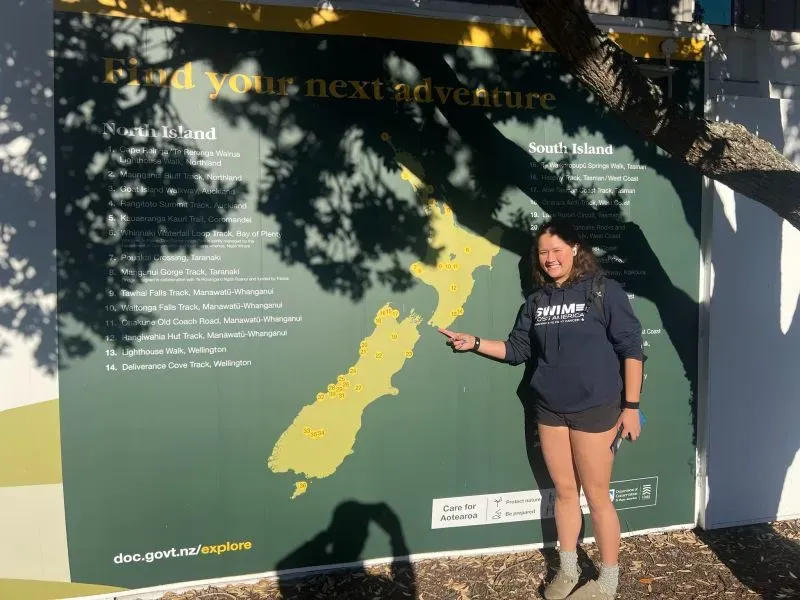 فتاة تشير إلى خريطة نيوزيلندا، محاطة بأشجار، ترتدي سترة رياضية، تعكس شغفها بالمغامرات المائية.