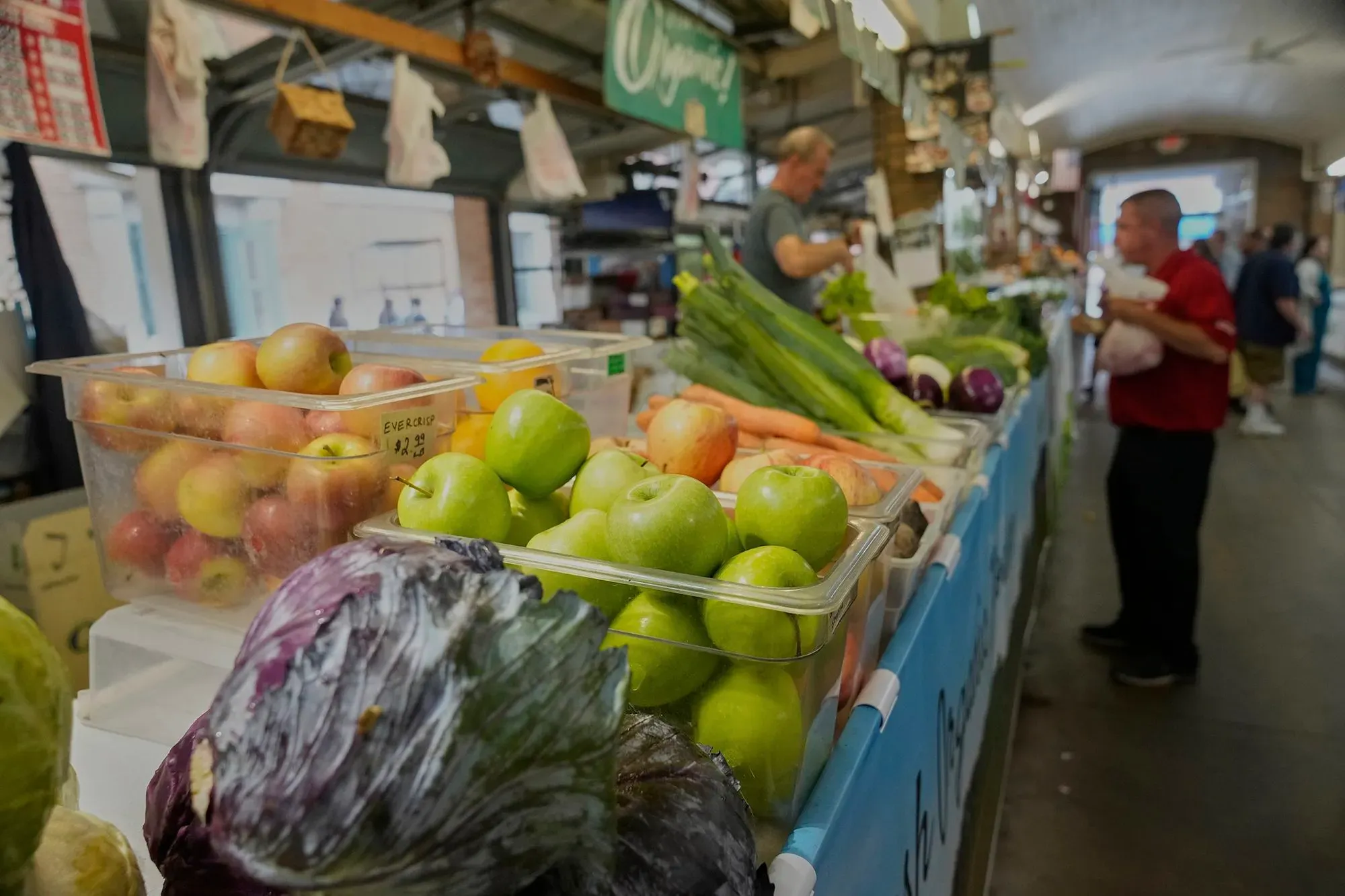 مشهد لسوق محلي حيث تُعرض الفواكه والخضروات الطازجة، مع تواجد المتسوقين الذين يتفقدون الأسعار والمنتجات.