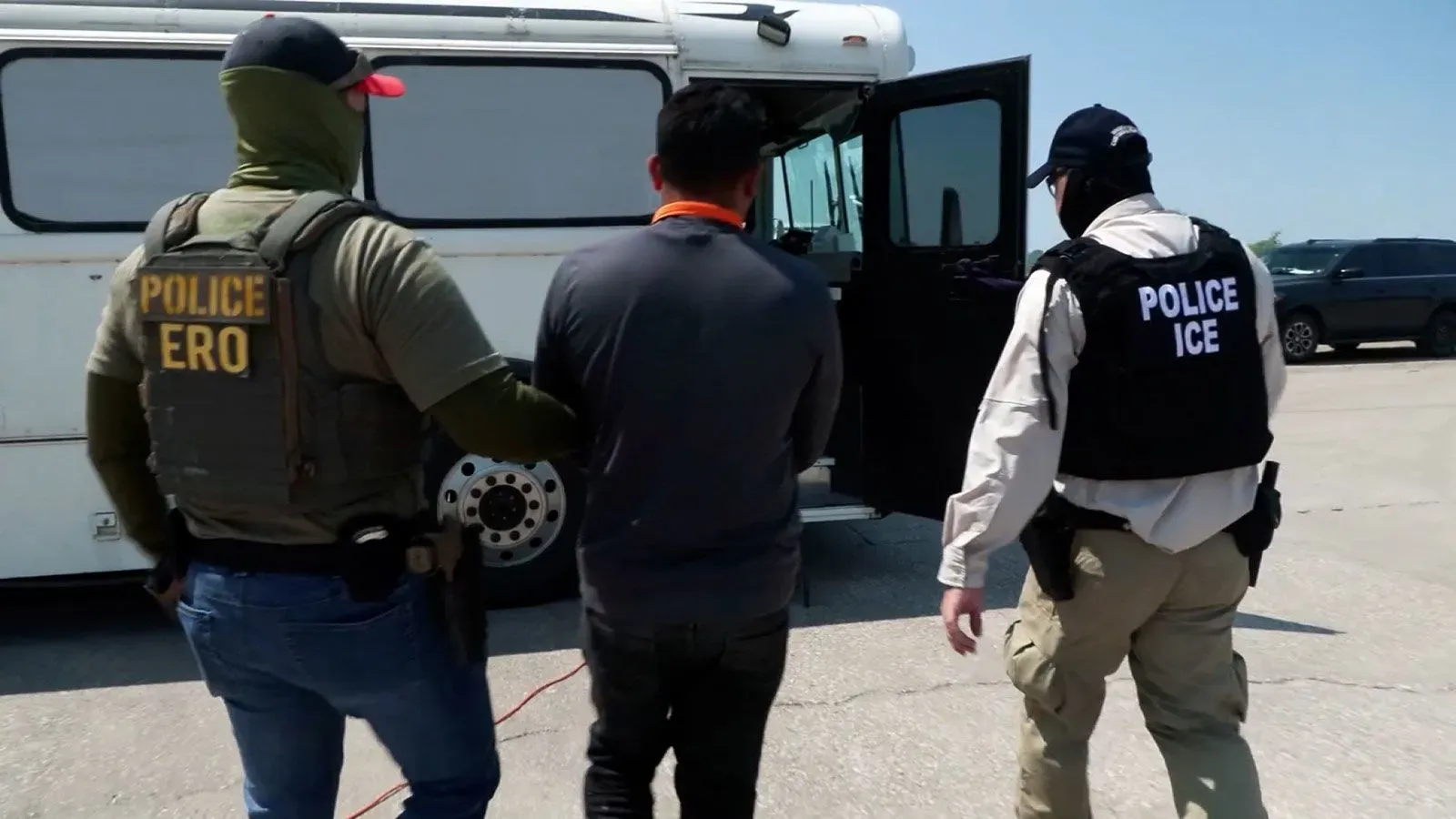 احتجاز مهاجر غير شرعي من قبل ضباط إنفاذ قوانين الهجرة، مع وجود حافلة للترحيل في الخلفية، يعكس سياسة الترحيل الحالية في الولايات المتحدة.