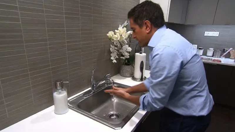 رجل يغسل يديه تحت الماء الجاري في مطبخ حديث، مع زهور صناعية وأدوات نظافة خلفه، مما يبرز أهمية غسل اليدين لصحة الفرد.