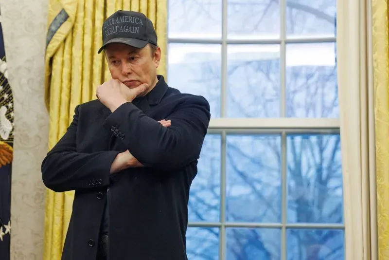 إيلون ماسك، مرتديًا قبعة "اجعل أمريكا عظيمة مرة أخرى"، يتأمل بينما يقف في المكتب البيضاوي، مع نافذة خلفه.