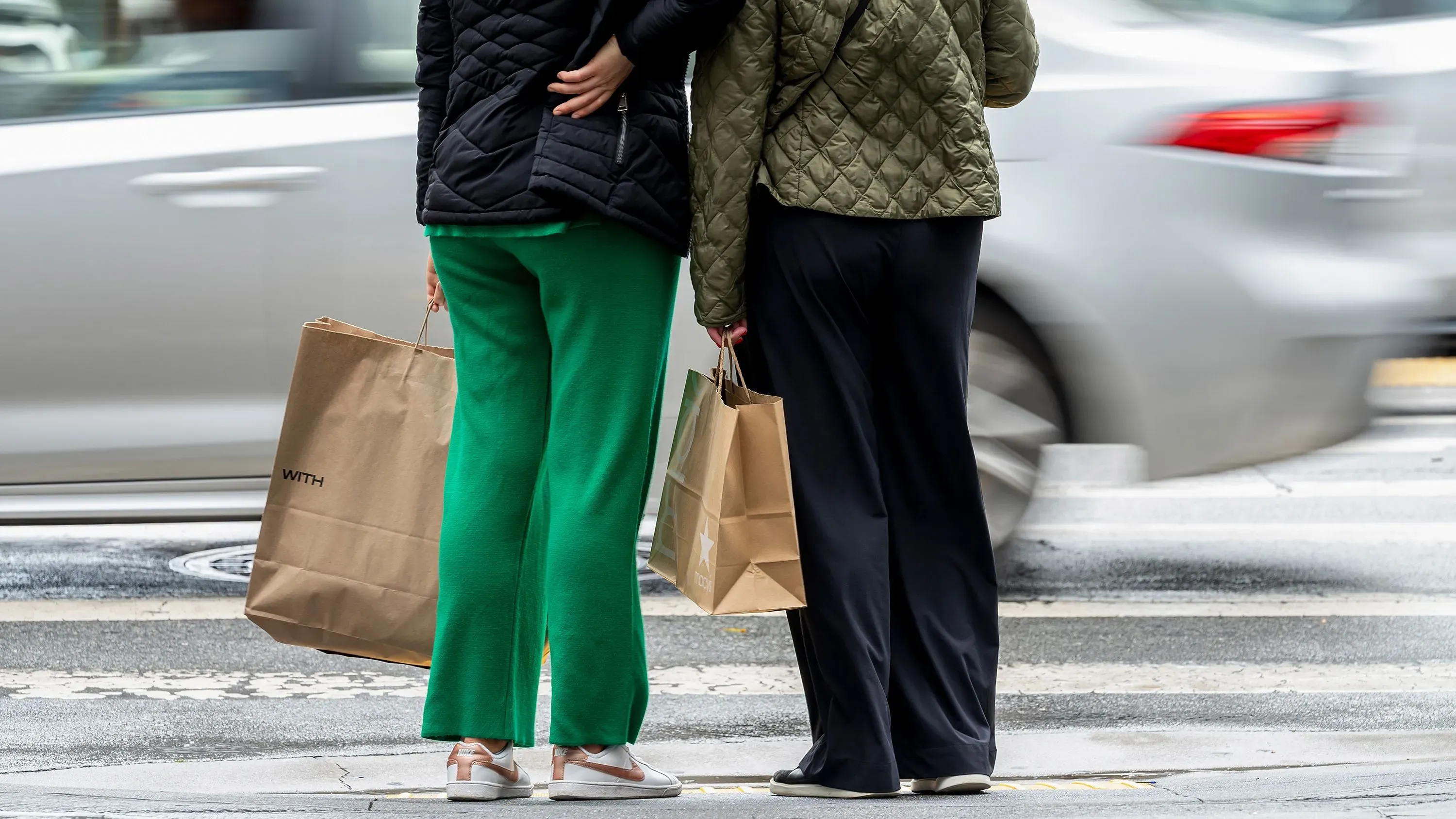 امرأتان تحملان أكياس تسوق، واقفتان على الرصيف، بينما تمر سيارة في الخلفية، مما يعكس تأثير الاقتصاد على سلوك المستهلكين.