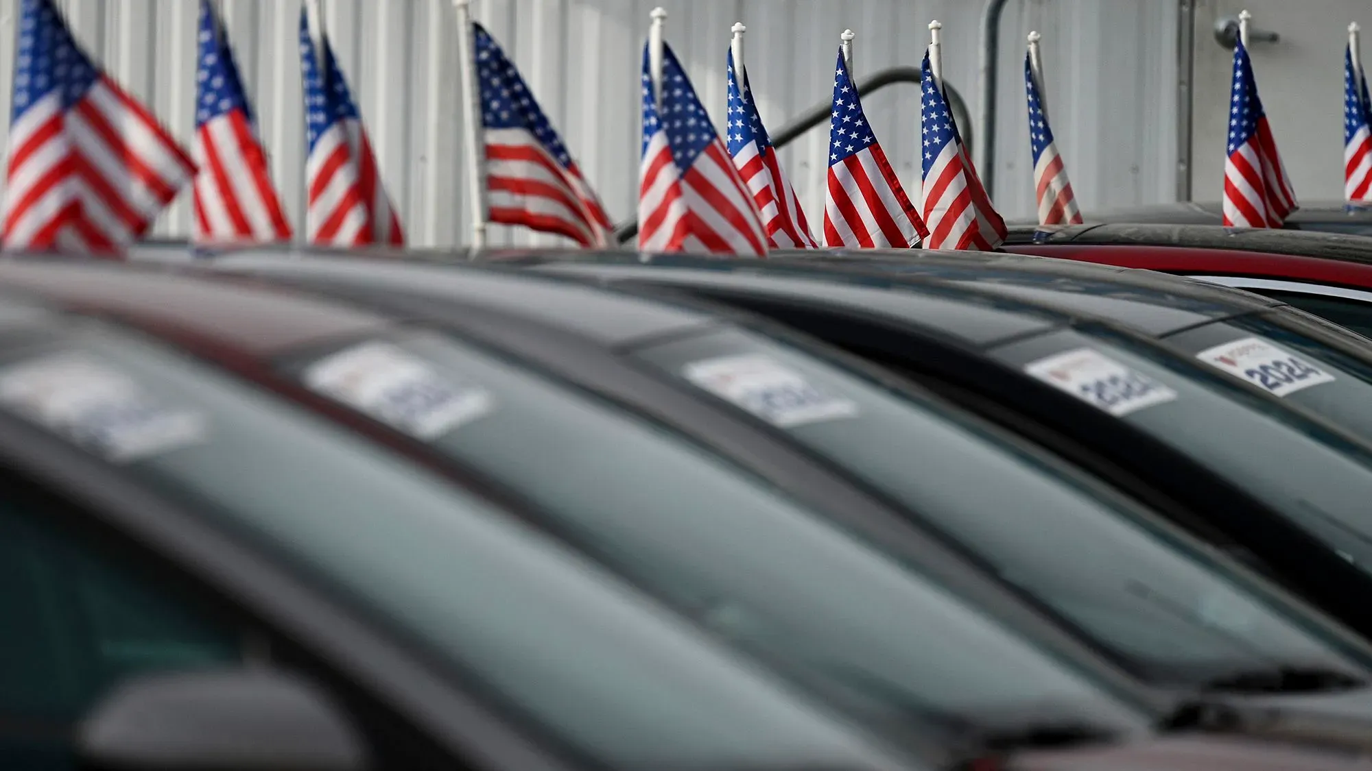 صفوف من السيارات مع أعلام أمريكية صغيرة ترفرف في معرض سيارات، تعكس تأثير التعريفات الجمركية على قرارات الشراء.
