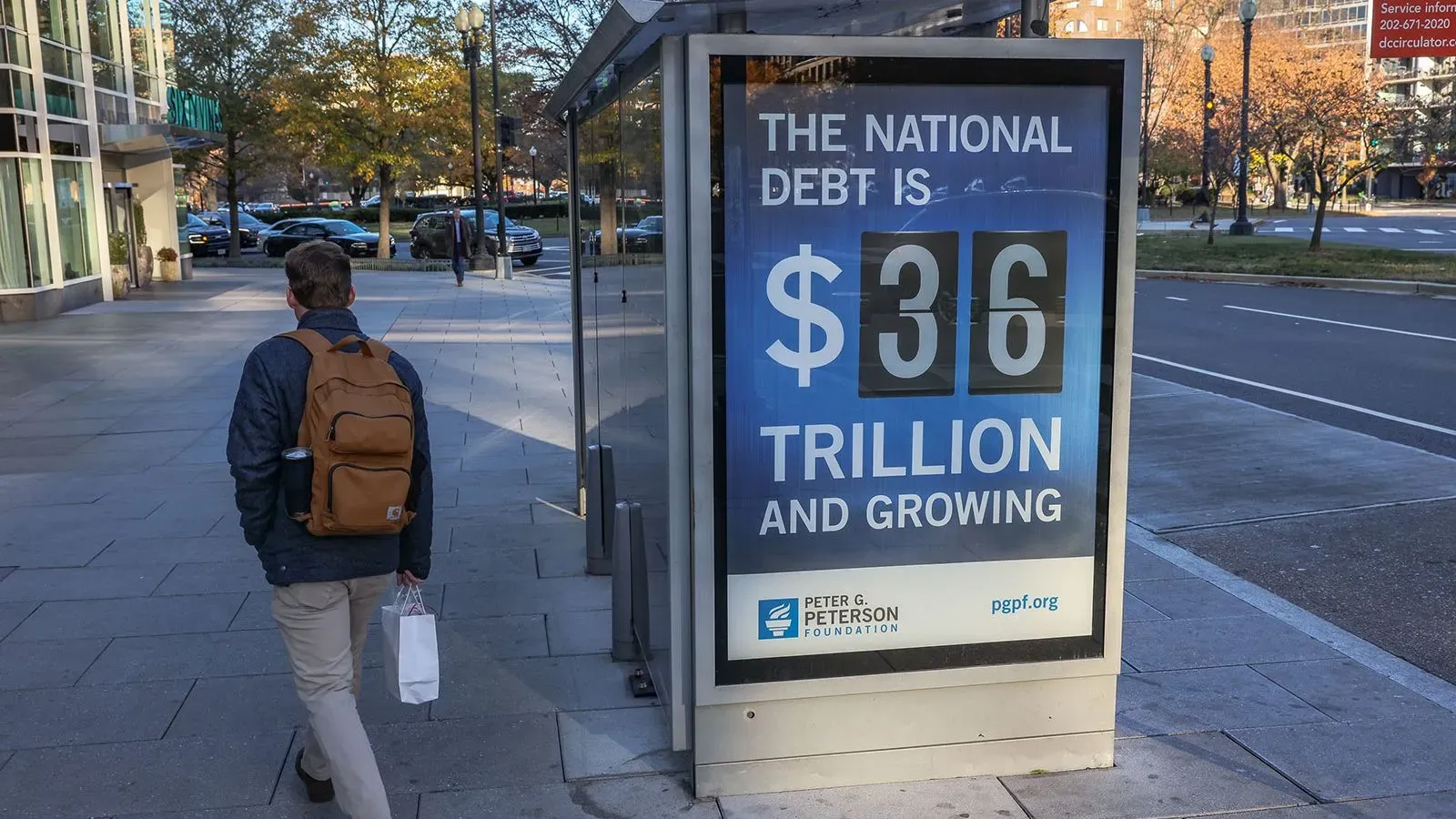 لافتة تعرض الرقم 36 تريليون دولار كديون وطنية أمريكية متزايدة، مع شخص يسير بجانبها في منطقة حضرية.