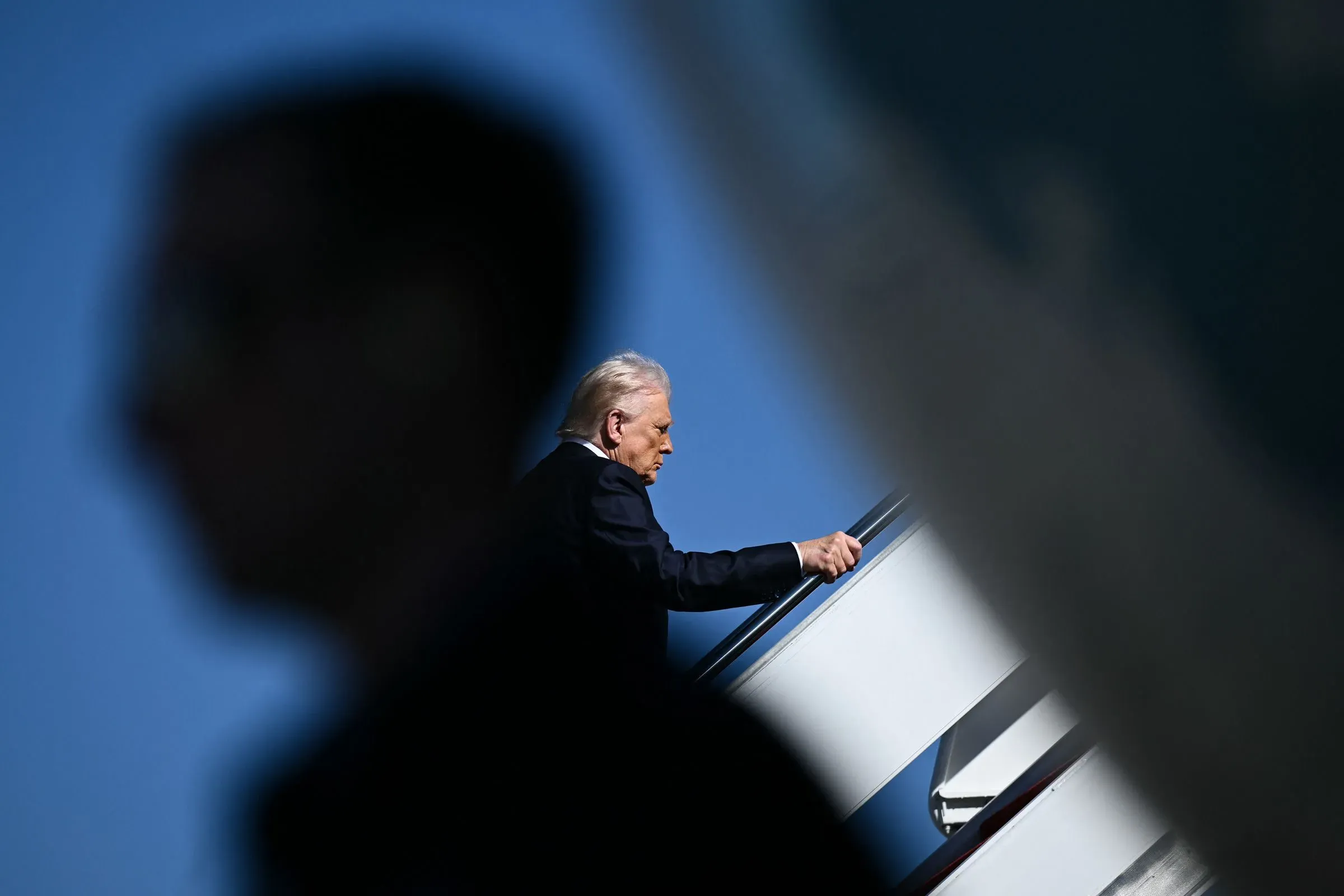 صورة لرجل يصعد على درج طائرة، يظهر في الخلفية ظلال لأشخاص آخرين. تعكس الصورة التوتر السياسي في الولايات المتحدة.