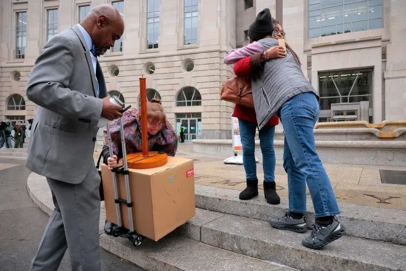 مشهد يظهر ثلاث نساء يتعانقن خارج مبنى حكومي، بينما يحمل رجل ببدلة علبة كبيرة على عربة. يبرز التفاعل العاطفي بين النساء.