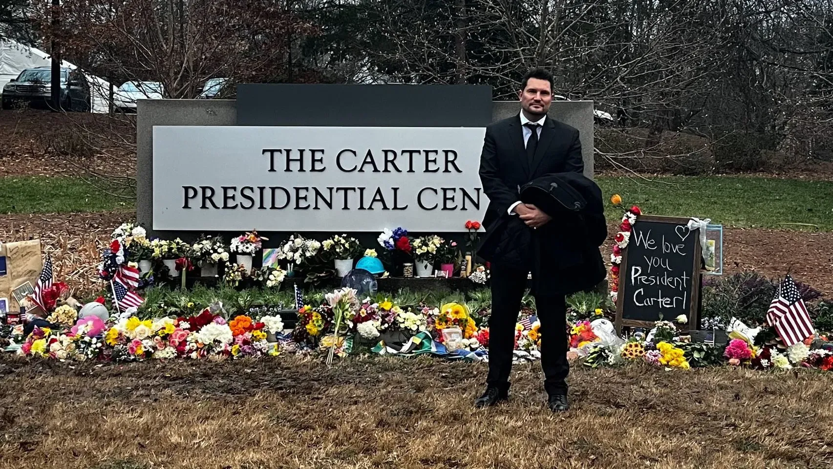 رجل يقف أمام مركز كارتر الرئاسي، محاطًا بالزهور والرسائل التذكارية، تعبيرًا عن الحب والاحترام للرئيس الأسبق جيمي كارتر.