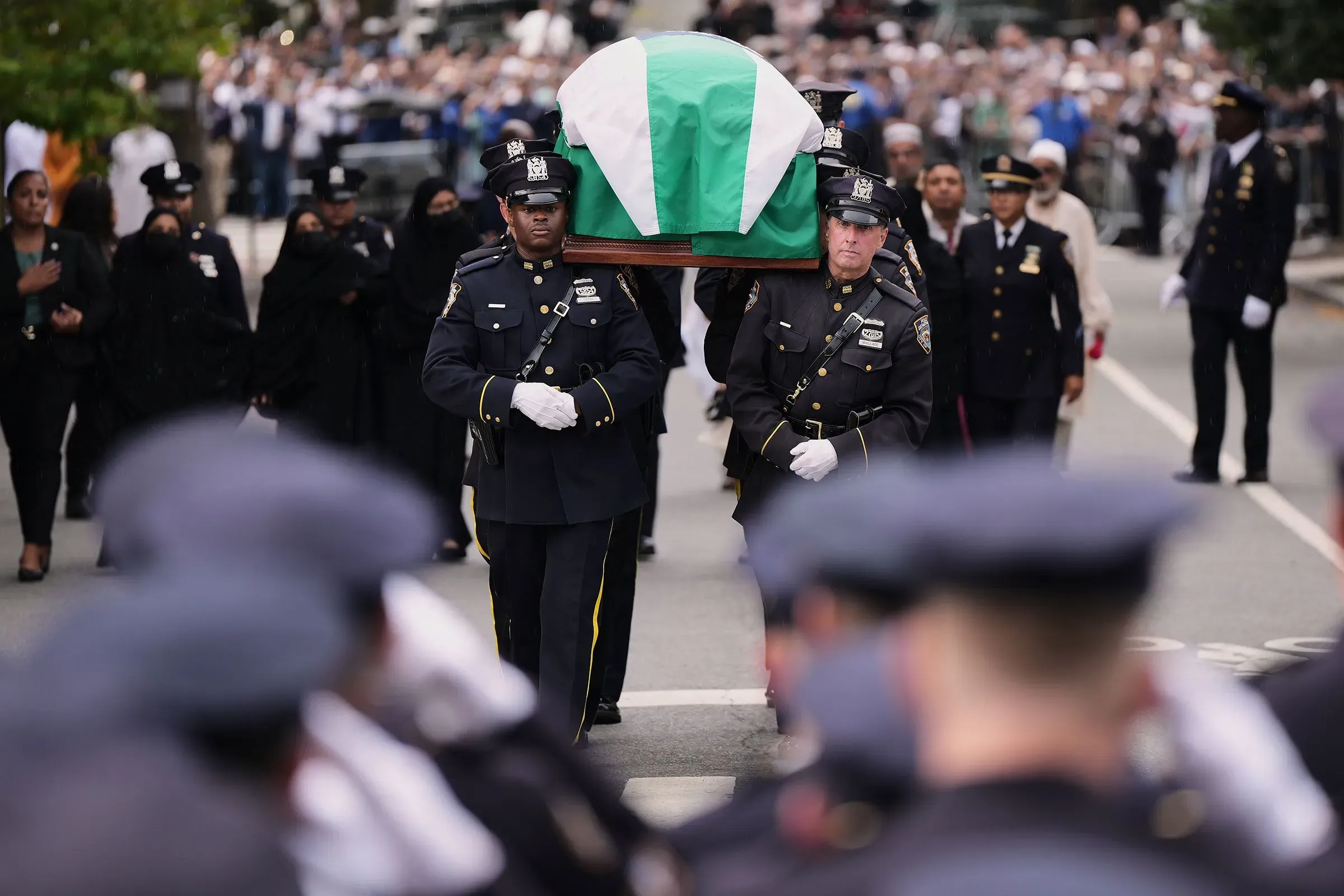 موكب جنازة ضابط شرطة يحمل النعش المغطى بعلم، مع حضور كبير من رجال الشرطة والمواطنين في الشارع، يعكس أجواء الحزن والتضامن.