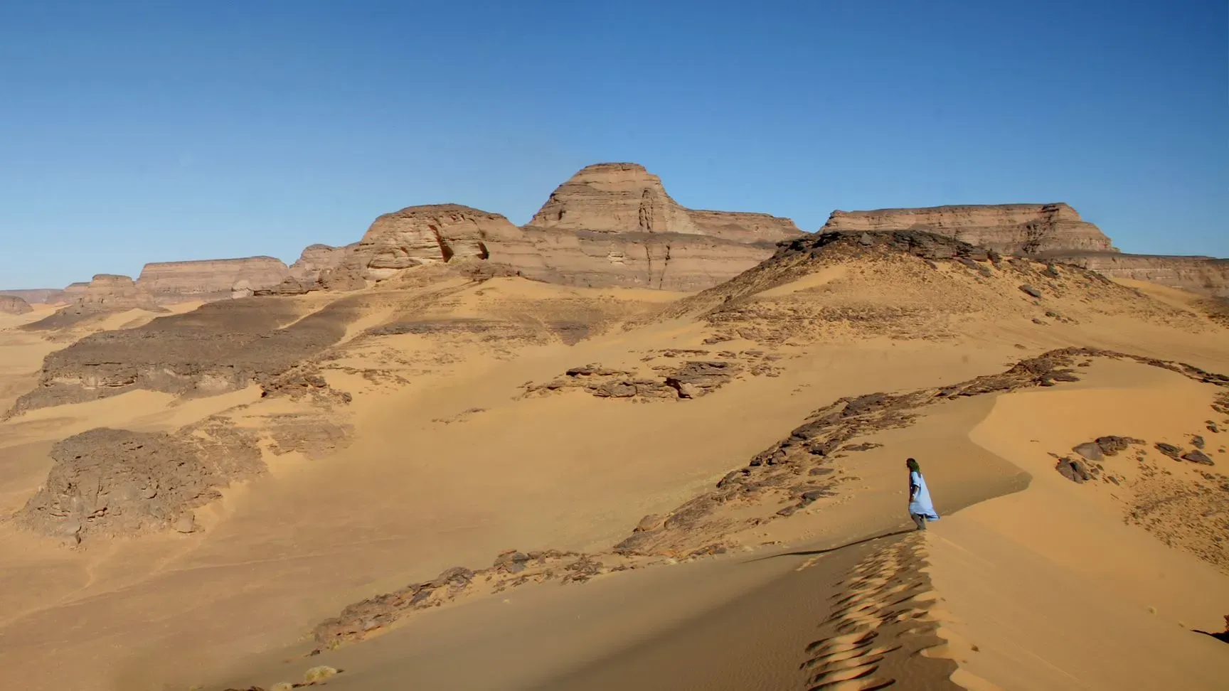 منظر طبيعي لصخور تاكاركوري في ليبيا، حيث تتداخل الكثبان الرملية مع التضاريس الجرداء، مما يعكس تاريخ المنطقة الغني.