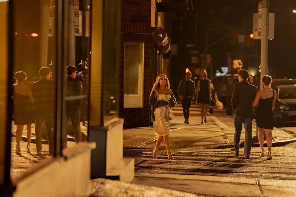 مجموعة من الأشخاص يسيرون في شارع مزدحم ليلاً، مع امرأة ترتدي فستاناً أنيقاً تحمل حقيبة، تعكس الأضواء المحيطة أجواء المدينة.