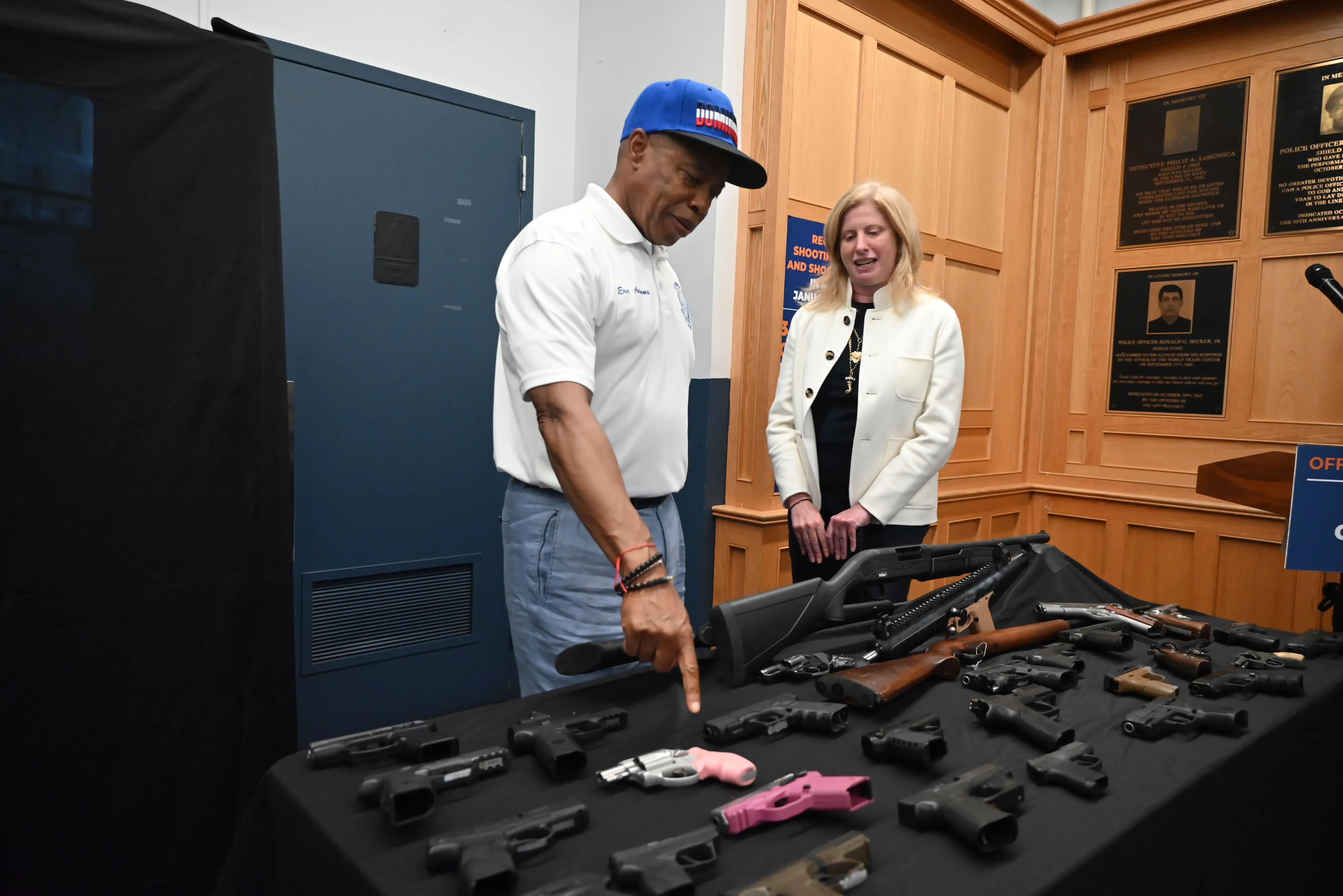رجل يشير إلى مجموعة من الأسلحة النارية على طاولة، بينما تقف امرأة بجانبه في مكان رسمي، مما يعكس قضايا الأمن والسلامة في نيويورك.