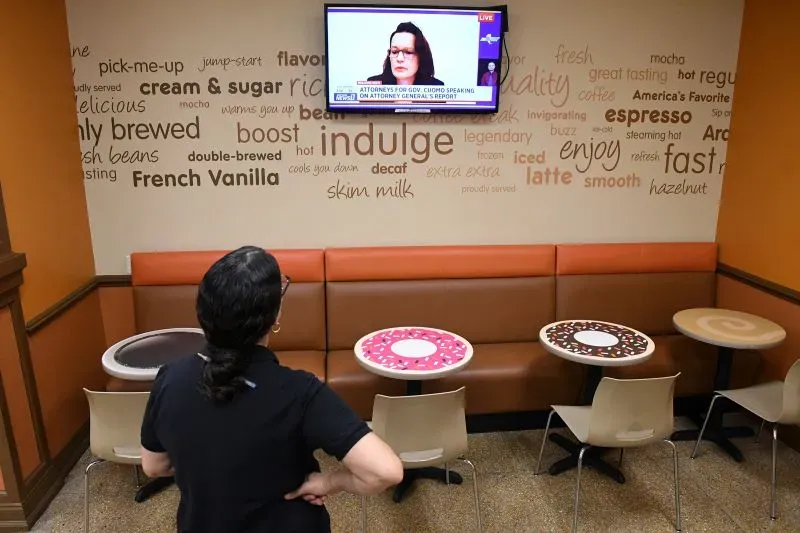 امرأة تشاهد تقريرًا إخباريًا على شاشة تلفاز في مقهى، حيث تتناول موضوع الشهادات ضد الحاكم السابق أندرو كومو.