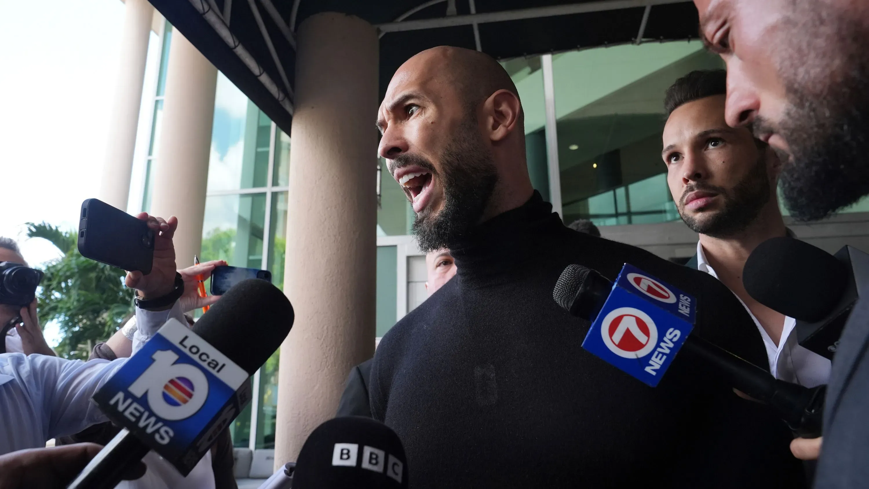 أندرو تيت يتحدث أمام الصحافة خارج المحكمة، محاطًا بالمراسلين، في خضم قضايا قانونية تتعلق بالاعتداء الجنسي والاتجار بالبشر.