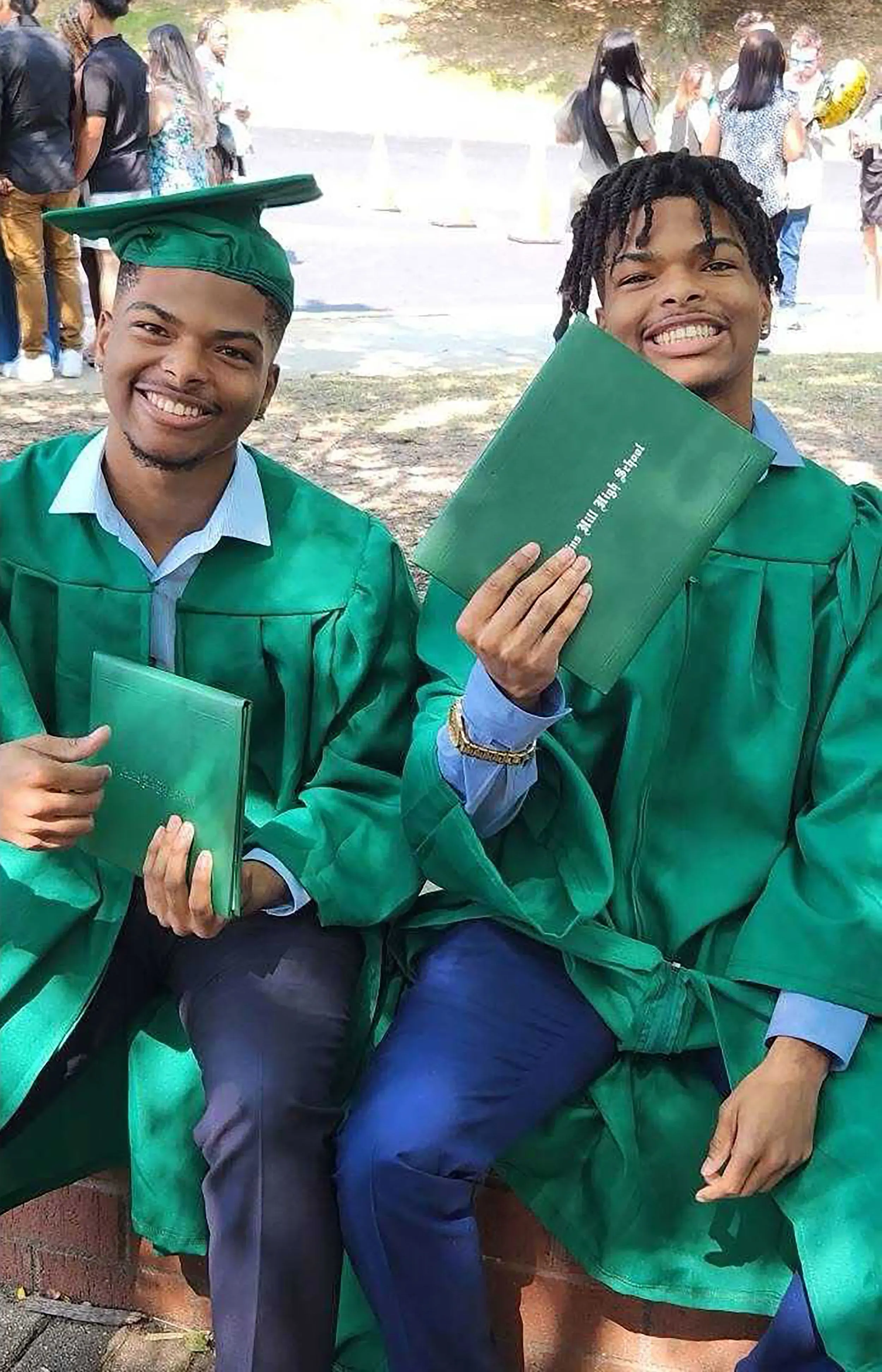 شقيقان توأمان يرتديان قبعات التخرج والعباءات الخضراء، يجلسان معاً مبتسمين ويحتفظان بشهادتيهما، في أجواء احتفالية.
