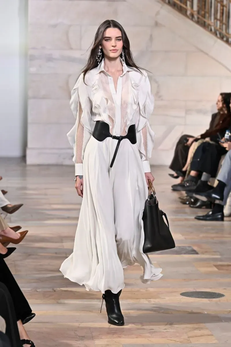 عارضة أزياء ترتدي فستانًا أبيضًا أنيقًا مع تفاصيل مبتكرة، تحمل حقيبة يد سوداء، أثناء عرض أزياء رالف لورين لخريف 2025 في معرض جاك شاينمان.