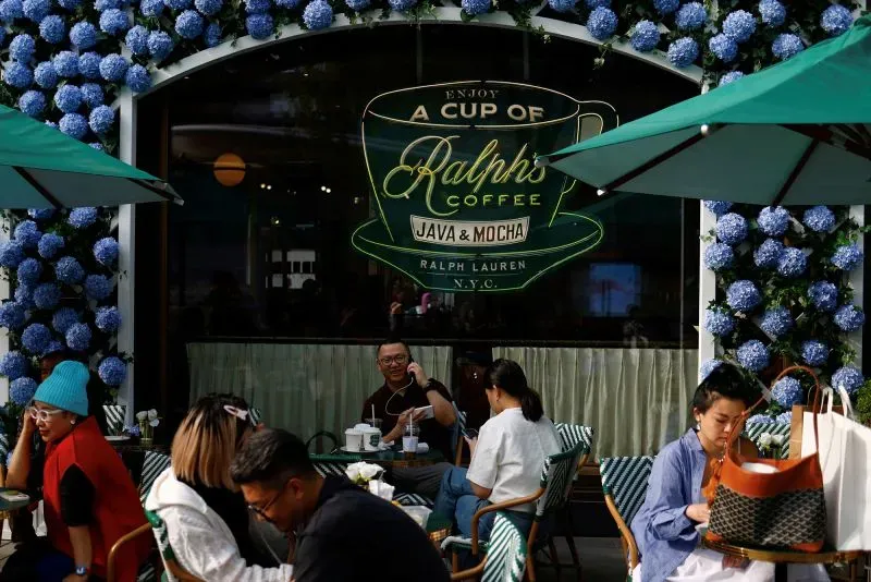 مشهد لمقهى "رالف لورين" مع زهور زرقاء على الواجهة، حيث يجلس الزبائن في الخارج ويتناولون القهوة، مما يعكس أجواء العلامة التجارية الأنيقة.