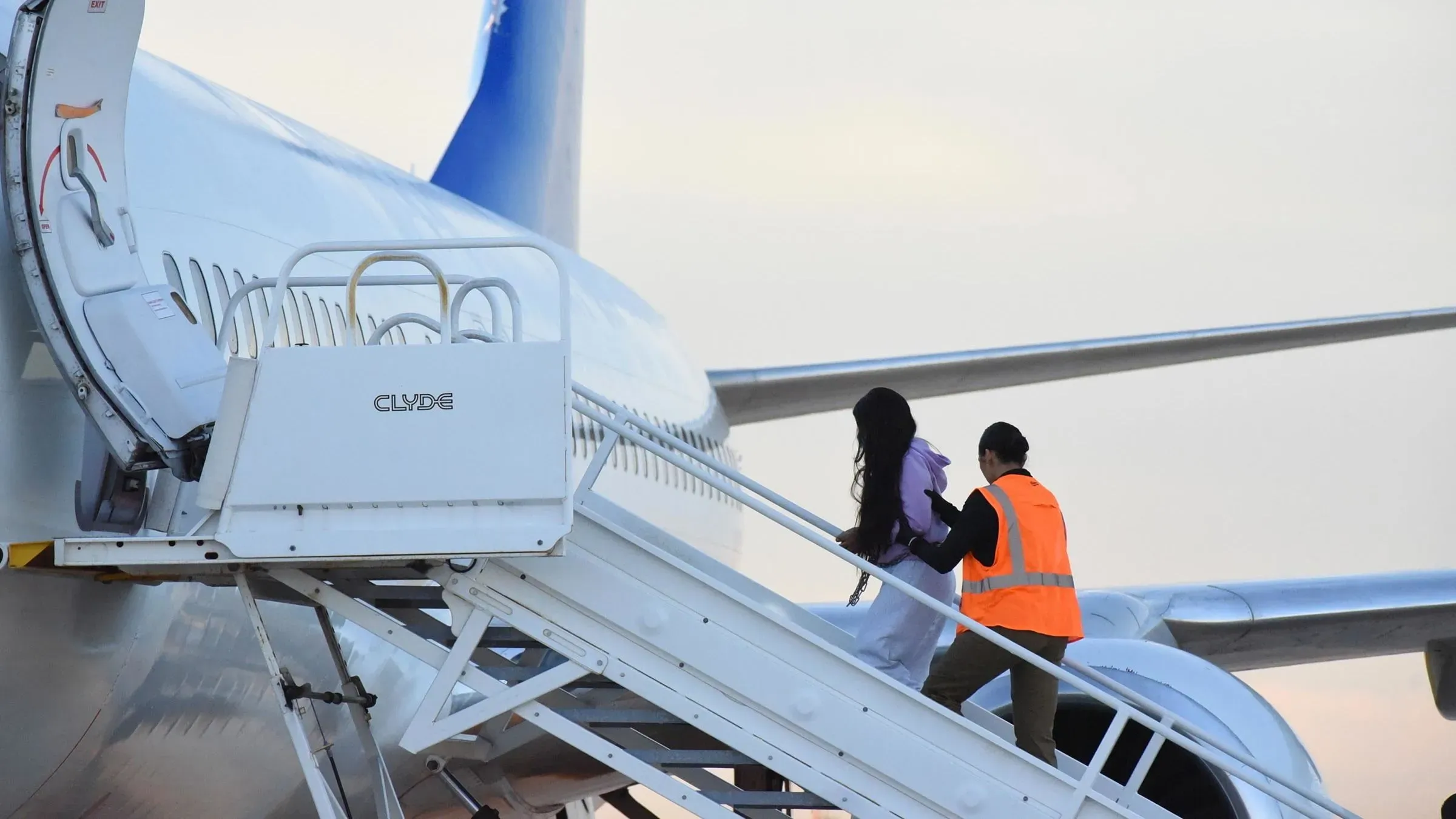 صورة لمهاجرين يتجهون إلى طائرة مع موظف يرتدي سترة برتقالية، تعكس إجراءات الترحيل القسري في ظل قوانين الهجرة.