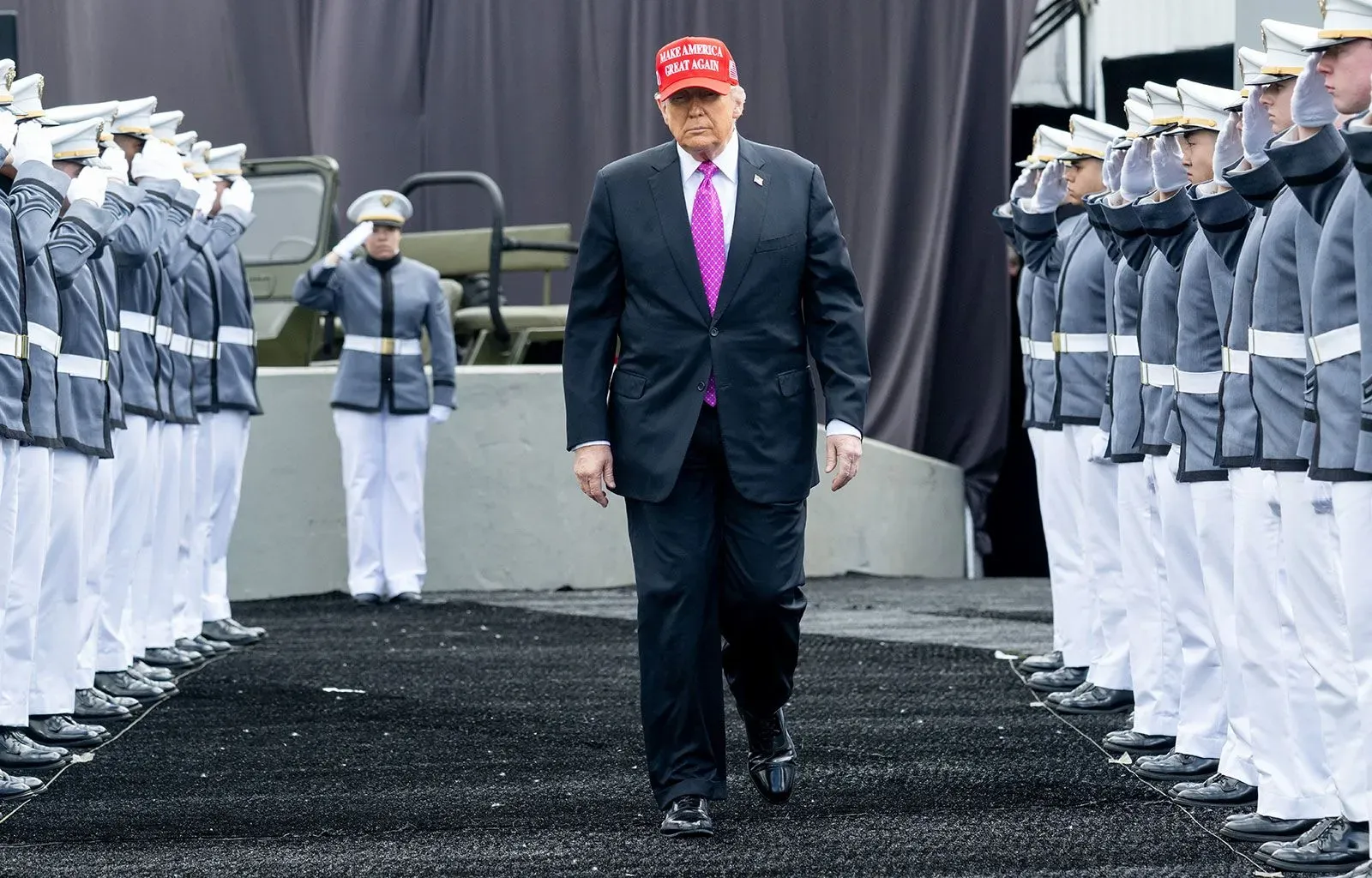 ترامب يسير بين صفوف من العسكريين في الأكاديمية العسكرية، مرتديًا قبعة تشير إلى حملته الانتخابية، مع خلفية عسكرية.