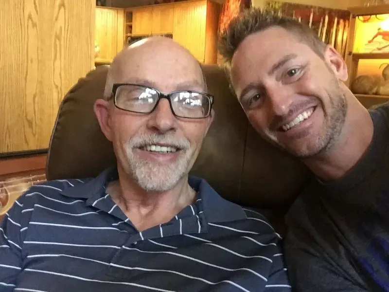 صورة لرجل مسن يرتدي نظارات ويبتسم بجانب رجل شاب مبتسم، يجلسان معًا في غرفة مريحة. تعكس الصورة روح الأمل في أبحاث الزهايمر.