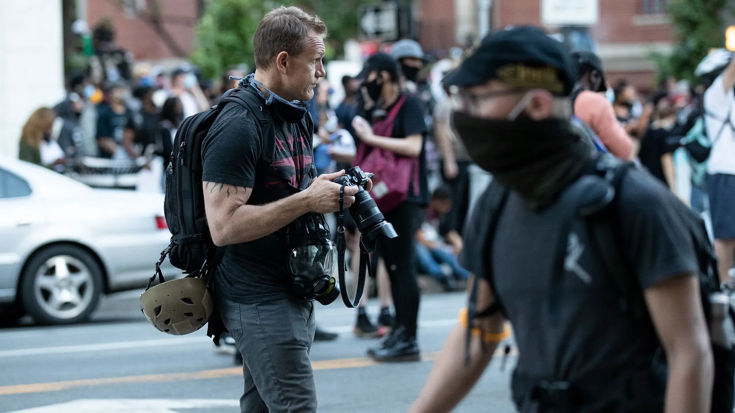 مصور محترف يحمل كاميرا ويتجول بين حشد من المتظاهرين، يعكس التوترات في تغطية الأحداث السياسية.