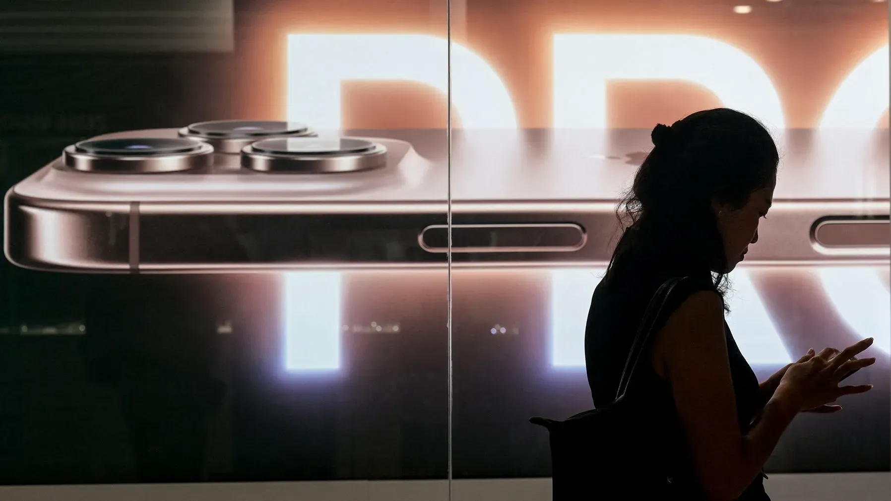 امرأة تتصفح هاتفها الذكي أمام إعلان كبير لجهاز iPhone Pro، مع التركيز على الكاميرات الخلفية.