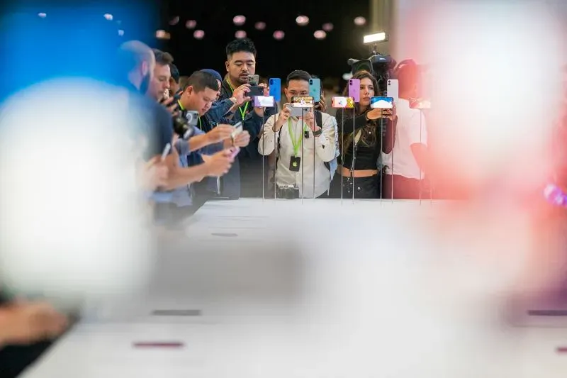 مجموعة من الأشخاص في حدث تقني، يلتقطون الصور باستخدام هواتفهم الذكية، مع وجود طاولة عرض في المقدمة.