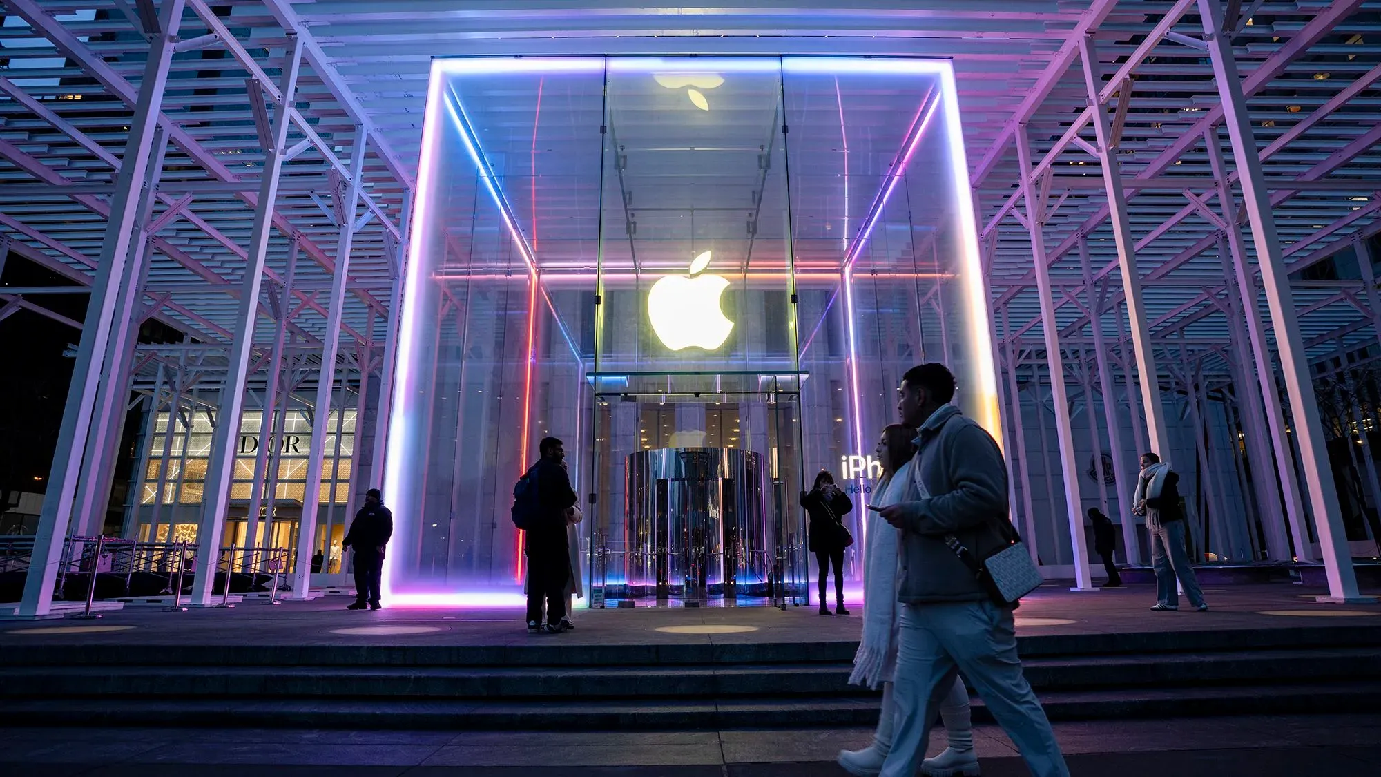 واجهة متجر آبل مضاءة بألوان زاهية، مع أشخاص يتجولون أمام المدخل الزجاجي، مما يعكس التوجه نحو الذكاء الاصطناعي وتحديثات سيري.