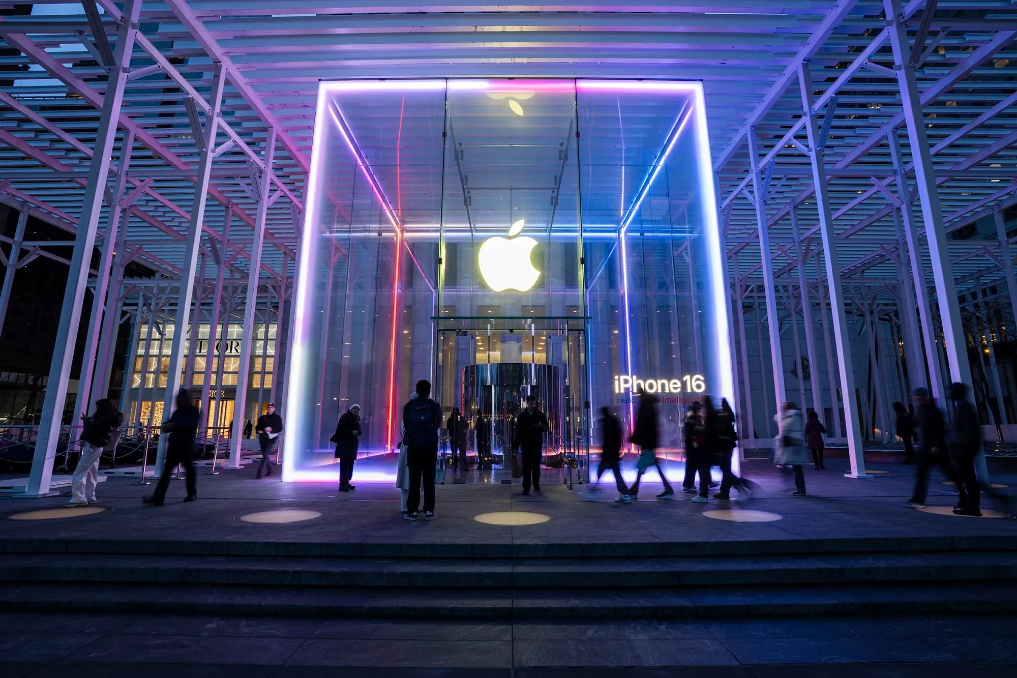 واجهة متجر آبل مضاءة بألوان زاهية، مع عرض لجهاز آيفون 16، والعديد من الزبائن يتجولون في الخارج، مما يعكس استثمار الشركة في الولايات المتحدة.