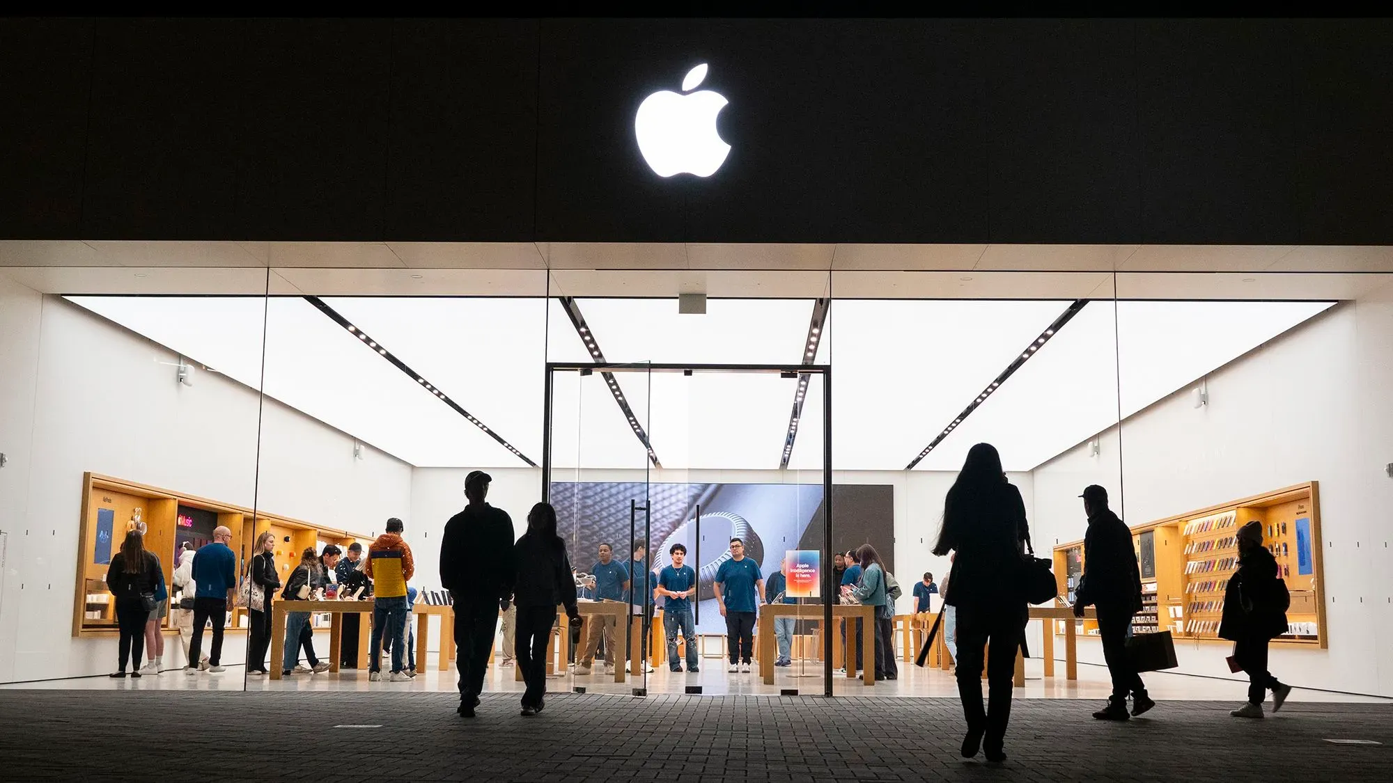 واجهة متجر آبل مضاءة، مع زبائن يتصفحون المنتجات داخل المتجر، وشعار آبل واضح في الأعلى، مما يعكس إطلاق iPhone 16e.