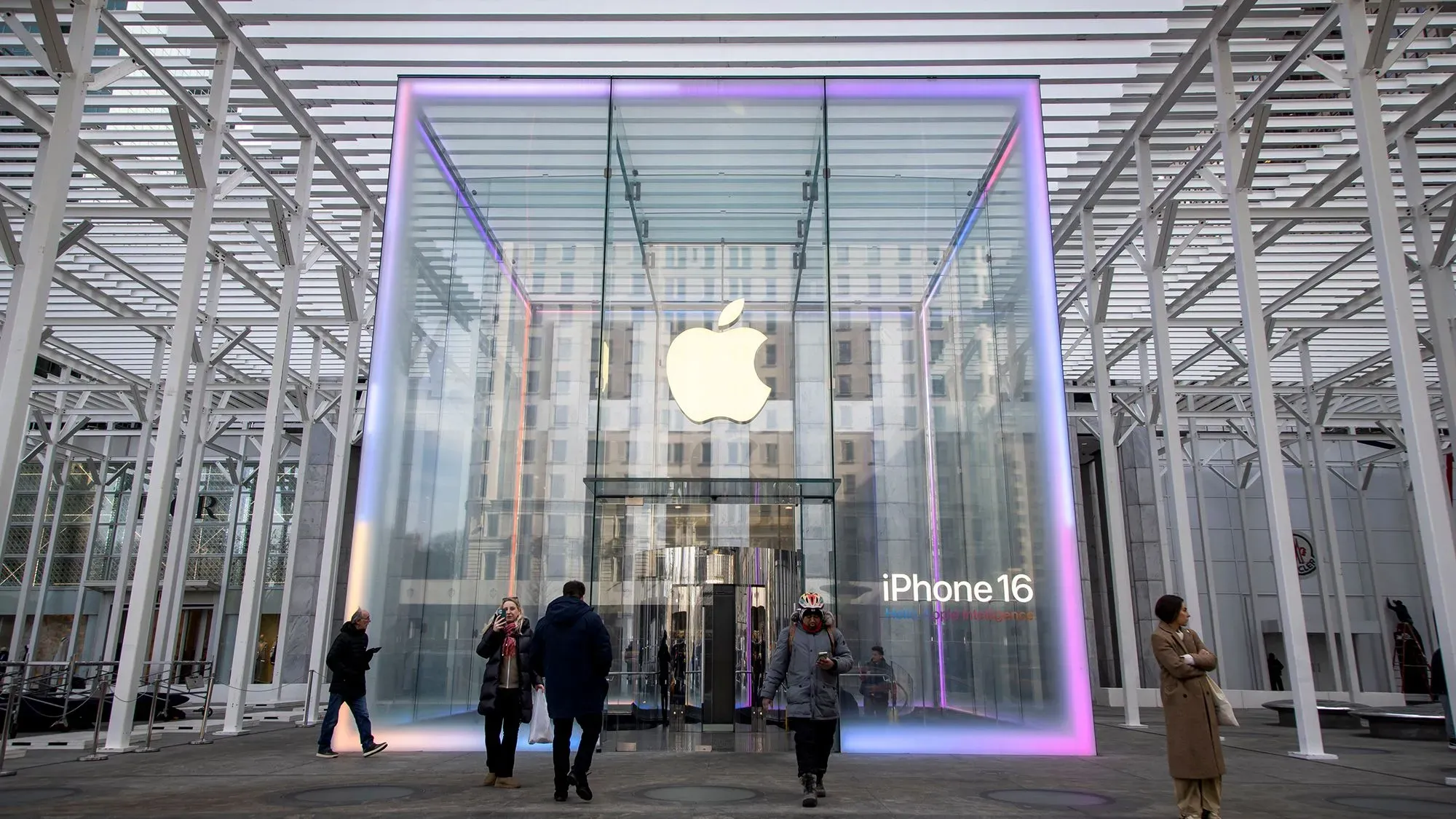 واجهة متجر آبل الزجاجية تعرض شعار الشركة وعبارة "آيفون 16"، مع زوار يتجولون حولها، تعكس الابتكار في عالم التكنولوجيا.