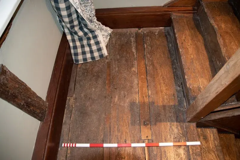 تفاصيل أرضية خشبية داخل منزل تاريخي، مع وجود شريط قياس، تشير إلى الموقع المحتمل لقصر هارولد غودوينسون في بوشام، إنجلترا.