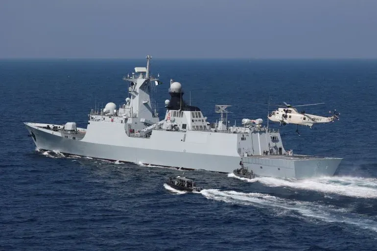 سفينة حربية تابعة للبحرية، ترافقها مروحية وقارب صغير، تبحر في المياه المفتوحة، تعكس الاستعدادات العسكرية والتوترات الإقليمية.