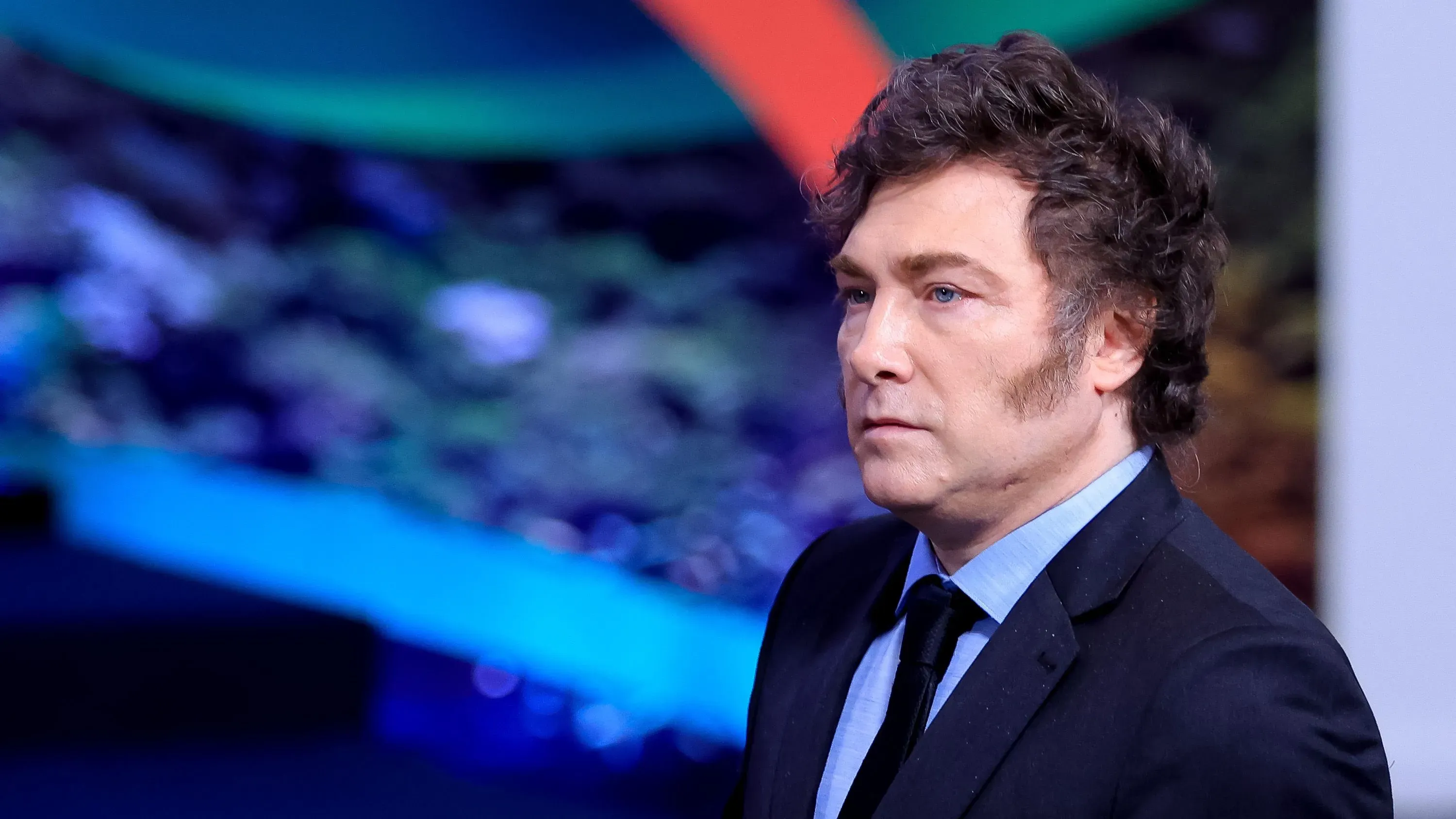 الرئيس الأرجنتيني خافيير ميلي يتحدث بجدية، محاطًا بألوان زاهية، وسط تداعيات فضيحة العملة المشفرة التي أثارت جدلًا واسعًا.