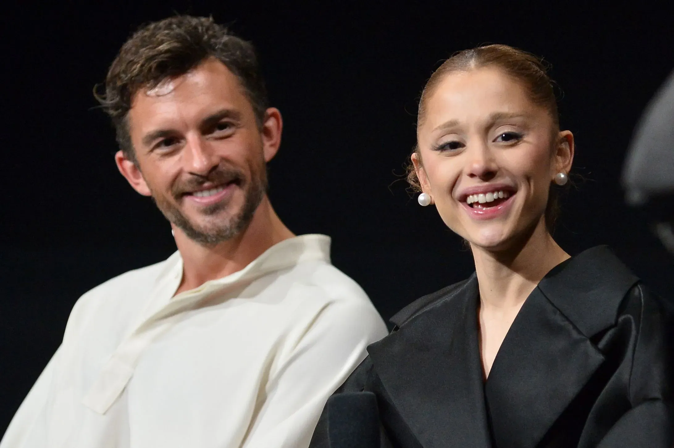 أريانا غراندي وجوناثان بيلي يبتسمان خلال حدث، مع خلفية داكنة، في إشارة إلى إحيائهما المسرحي "Sunday in the Park with George".