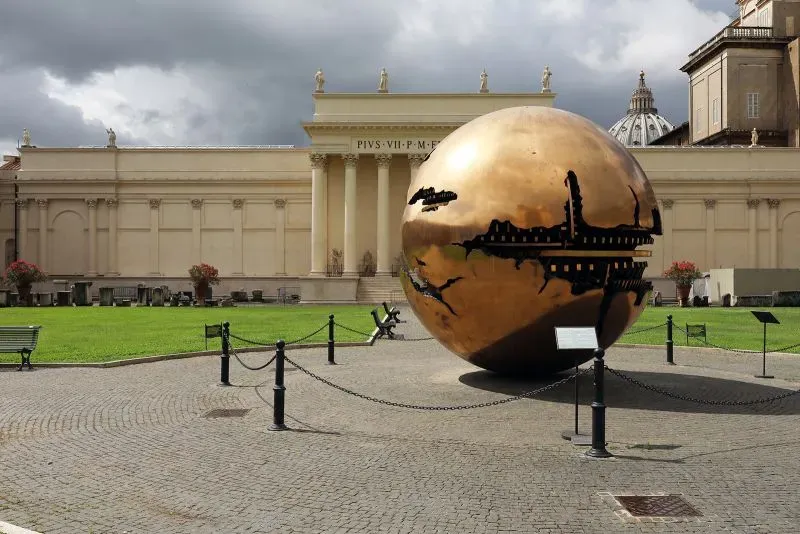 كرة برونزية ضخمة للفنان أرنالدو بومودورو، تقع في فناء متاحف الفاتيكان، تعكس تفاصيل معمارية محيطة وتظهر تصميمات داخلية معقدة.