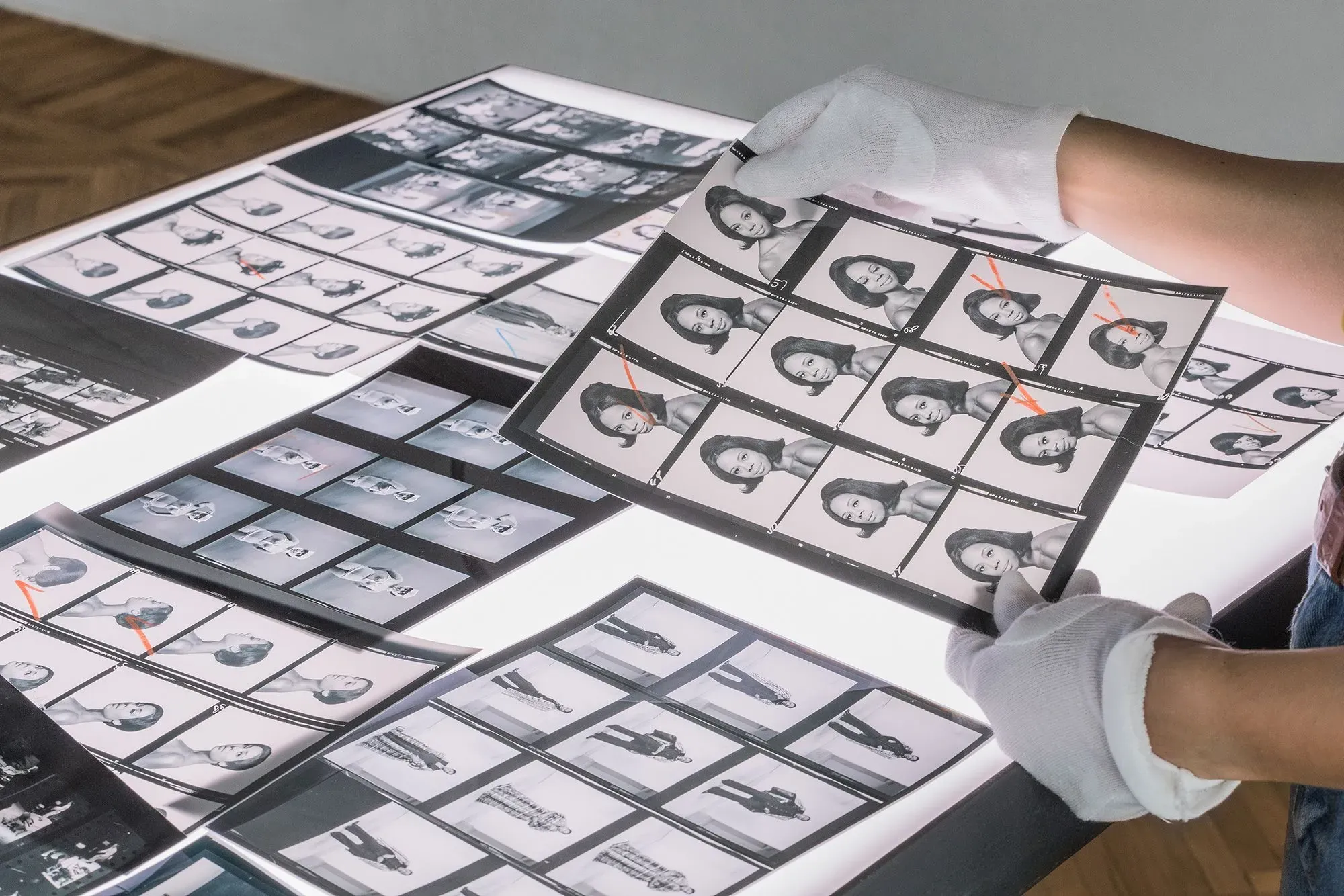 أيدٍ مرتدية قفازات بيضاء تراجع صورًا فوتوغرافية بالأبيض والأسود على طاولة مضاءة، تتضمن لقطات لنساء وأحداث تاريخية.