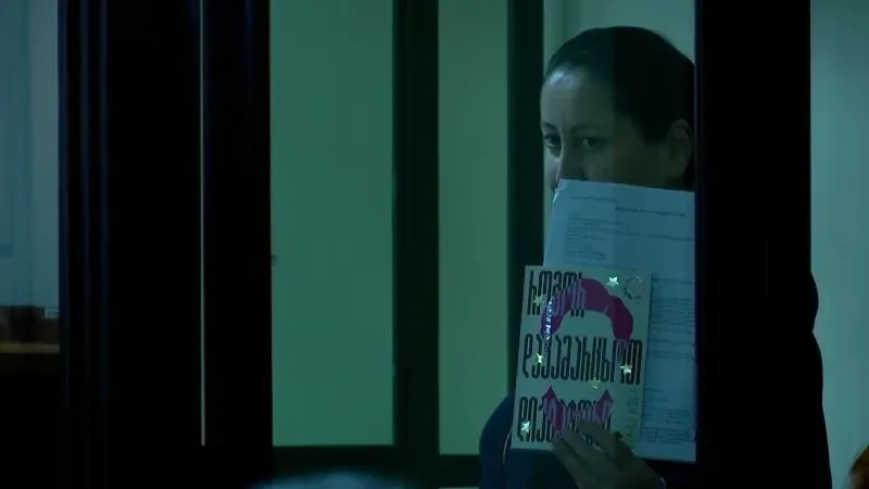 امرأة تحمل كتابًا وأوراقًا، تظهر من خلف نافذة زجاجية، تعكس أجواء التوتر السياسي في جورجيا.