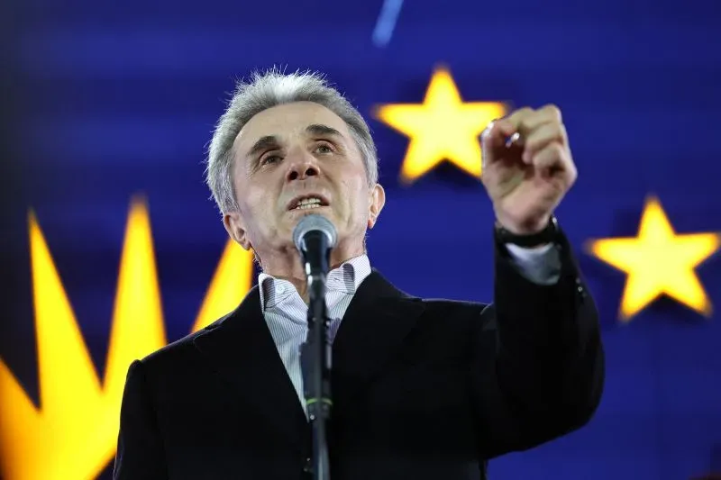 رجل يتحدث في تجمع عام، مع خلفية تحمل نجومًا زرقاء، يعبر عن موقفه تجاه الاحتجاجات في جورجيا.