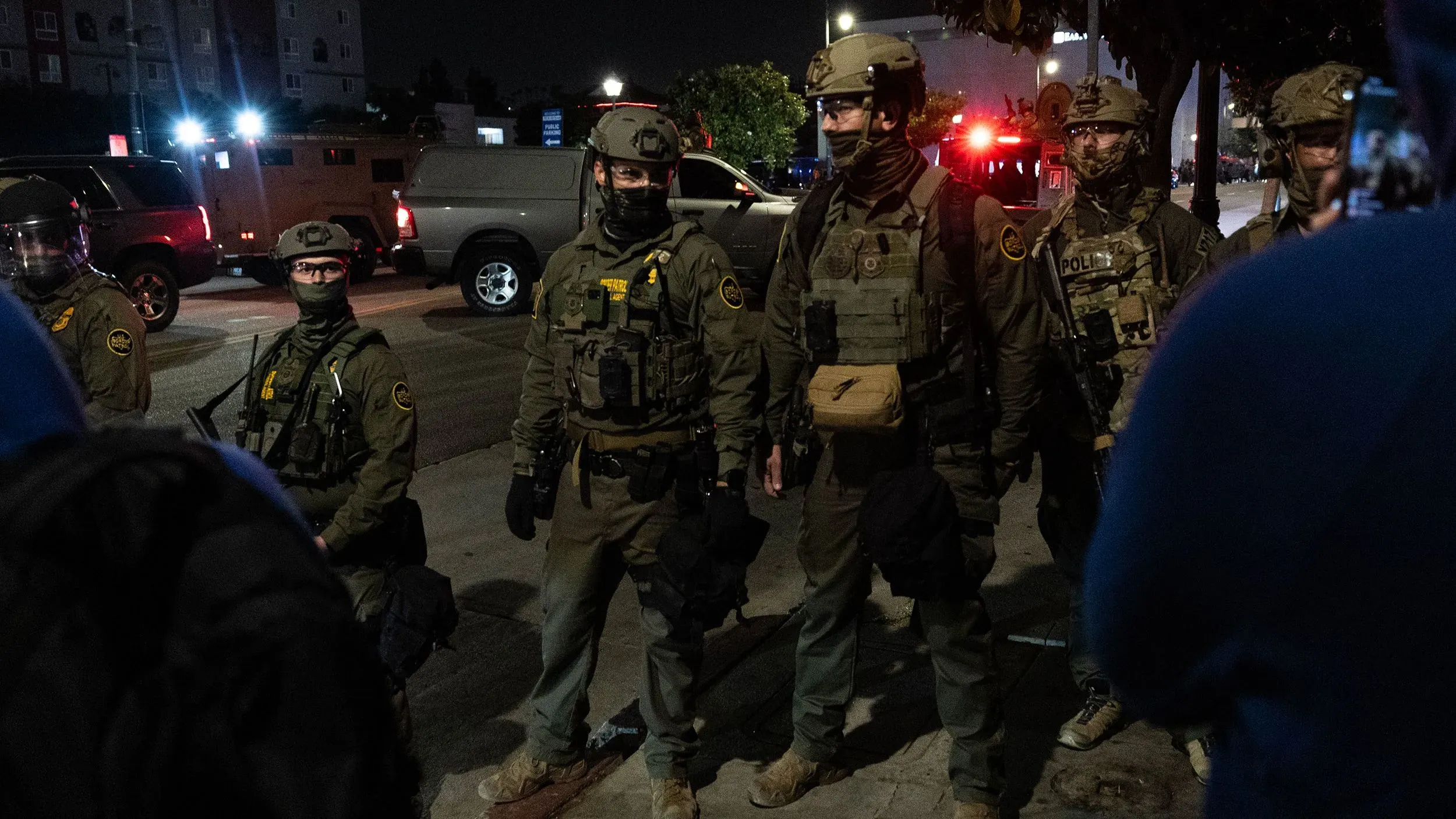 عناصر من الشرطة يرتدون زيًا عسكريًا خلال احتجاجات في لوس أنجلوس، مع وجود مركبات أمنية في الخلفية، تعكس التوترات حول مداهمات الهجرة.