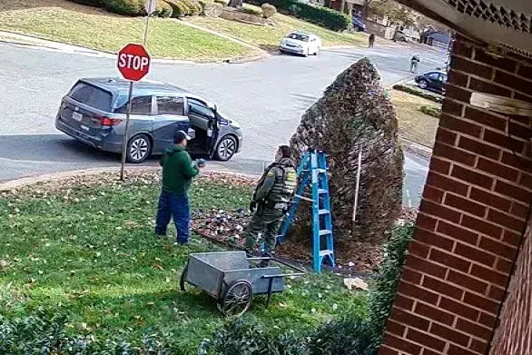 شاحنة صغيرة رمادية متوقفة بجانب عاملين في حديقة، أحدهما يرتدي زيًا عسكريًا، بينما يزينان شجرة بالأضواء في حي شارلوت.