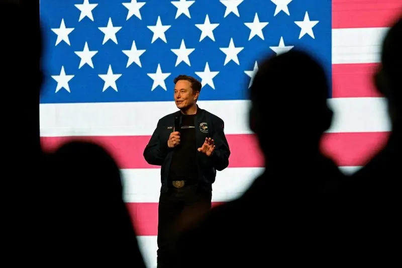 إيلون ماسك يتحدث أمام خلفية علم الولايات المتحدة، مع ظهور ظلال الحضور خلفه، في سياق حديثه عن إدارة الكفاءة الحكومية.