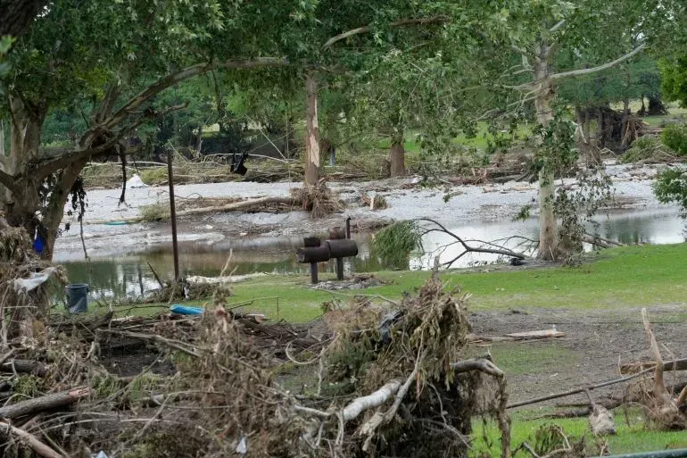 تظهر الصورة آثار الفيضانات في تكساس، حيث تتناثر الحطام والأشجار المكسورة، مع وجود مياه راكدة في الخلفية.